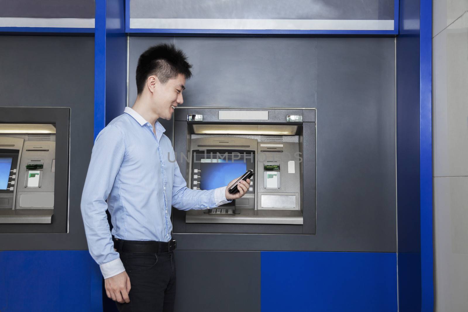 Smiling young businessman standing in front of an ATM and looking at his phone
