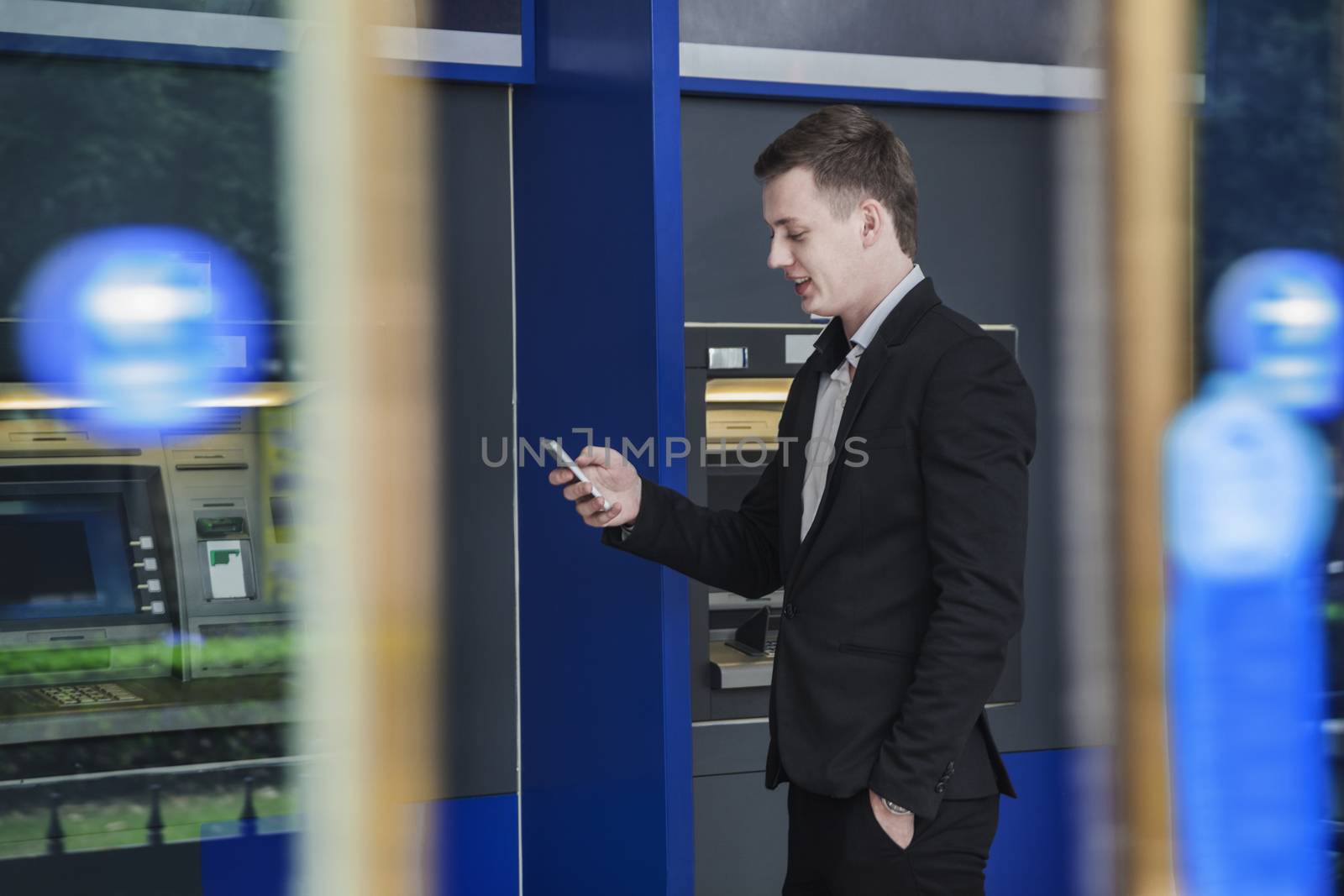 Smiling young businessman standing in front of an ATM and looking at his phone