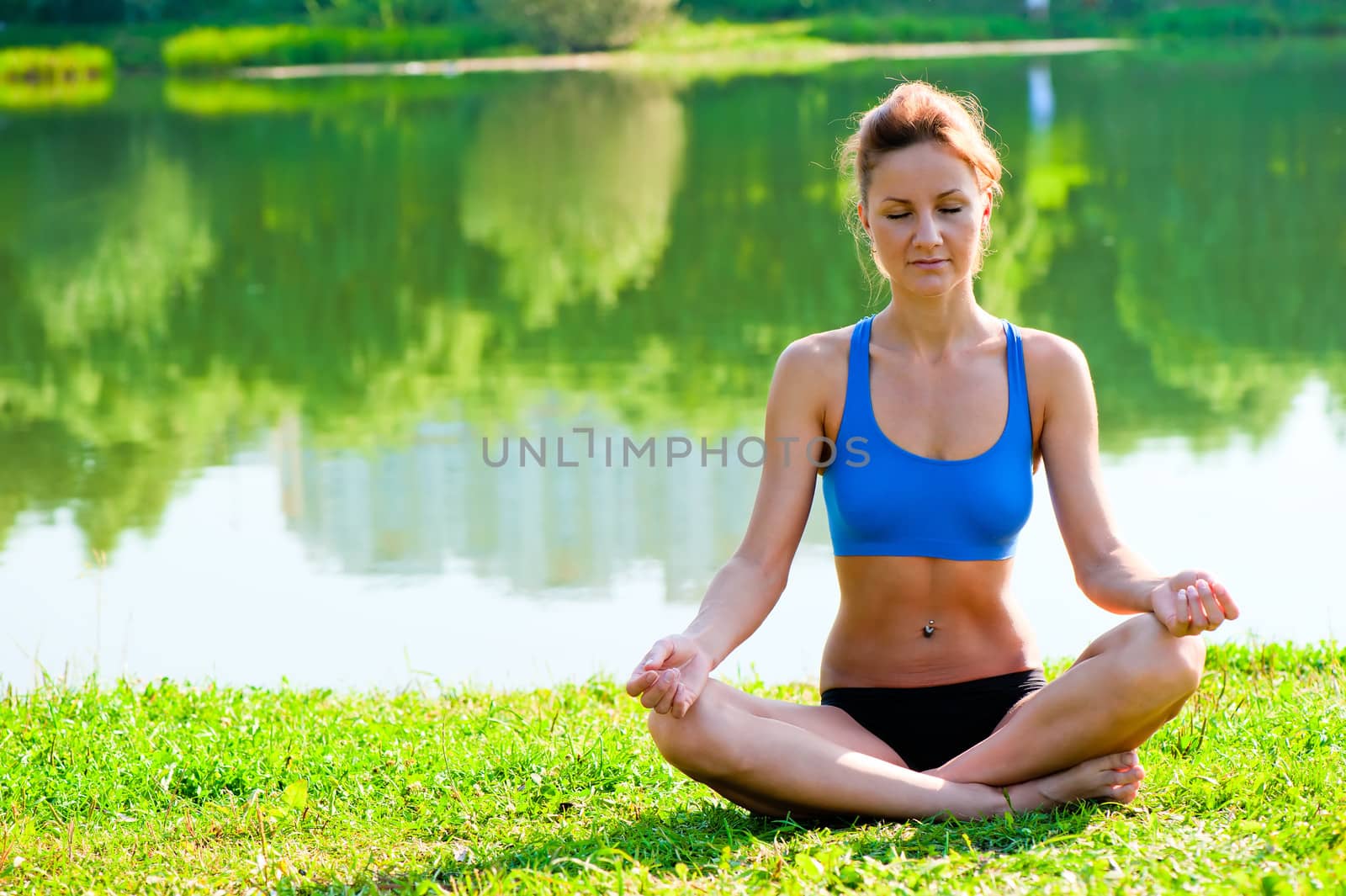 tightened girl in sportswear meditating in the lotus position at the lake in the park by kosmsos111