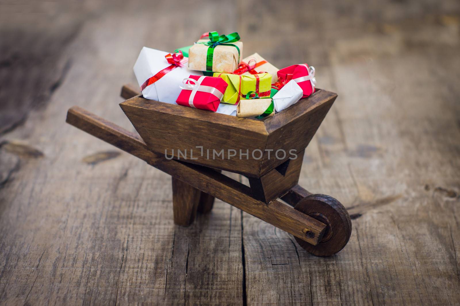 A wheelbarrow with many christmas presents on wood background.