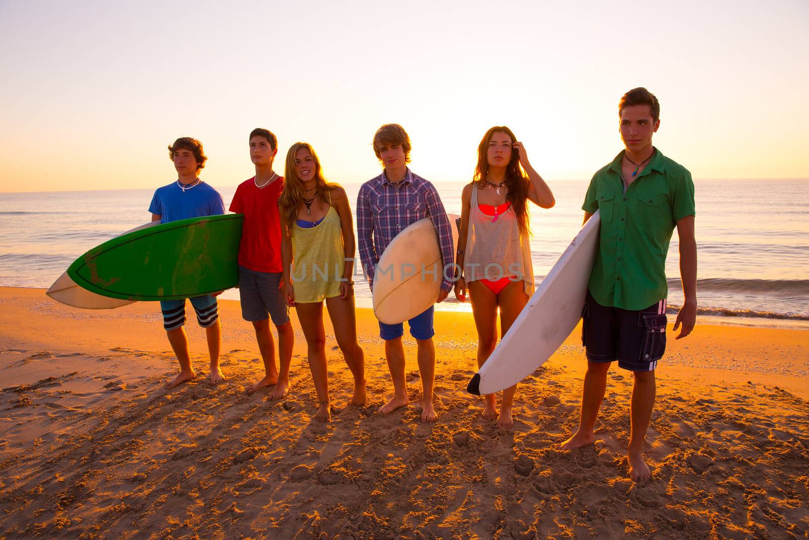 Surfers teen boys and girls group walking on beach at sunshine sunset backlight