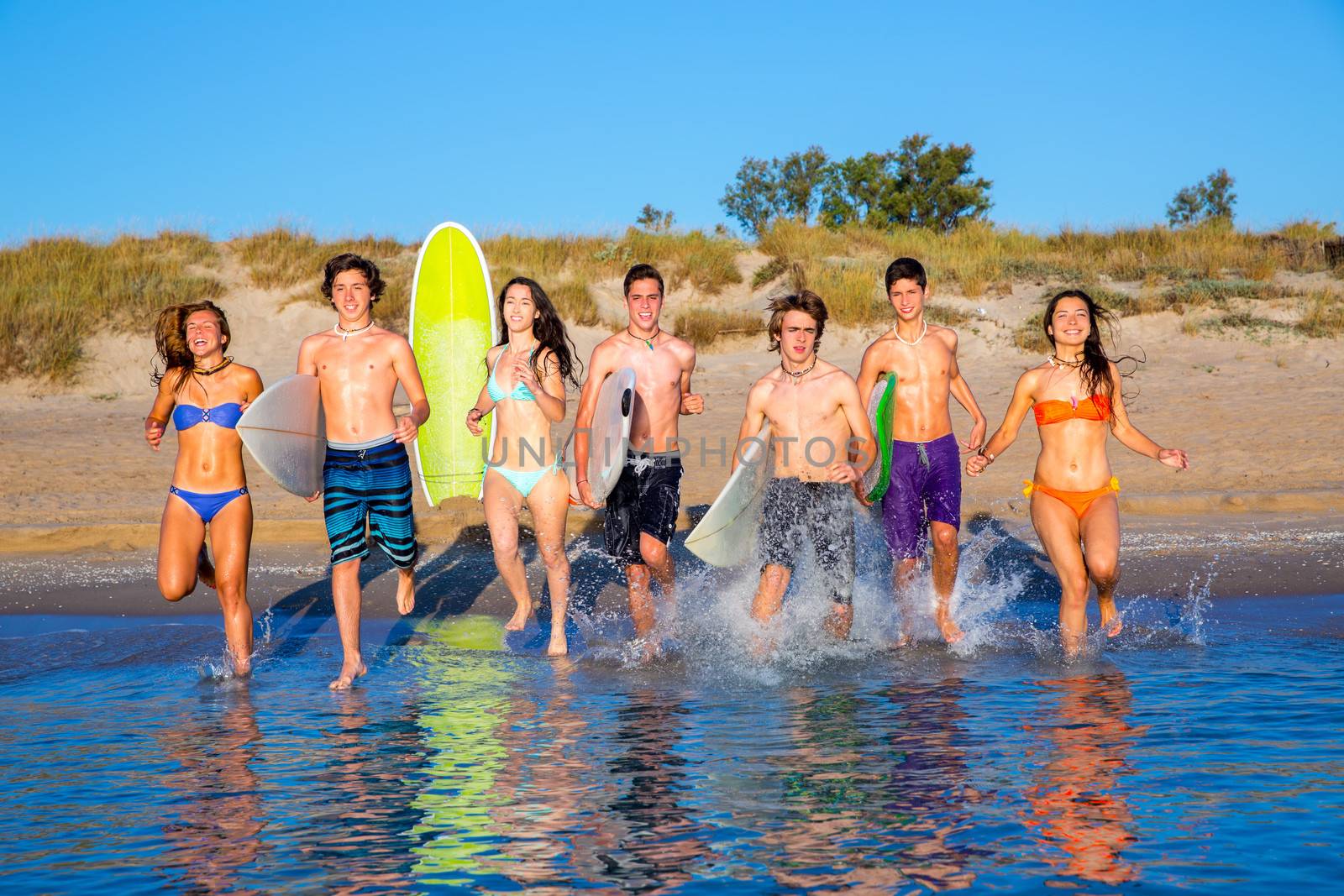 Teen surfers boys and girls group running happy to the beach splashing water