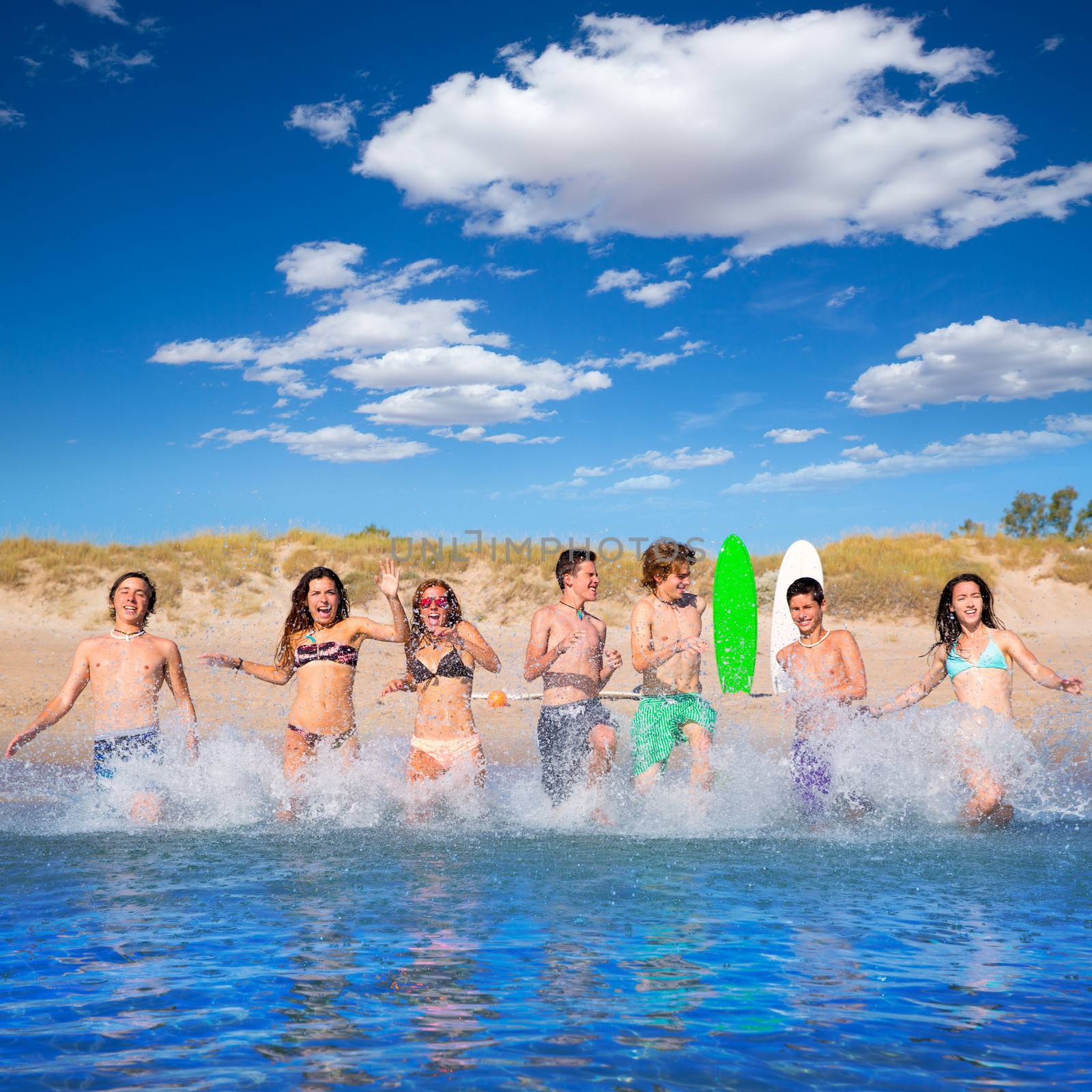 Teen surfers boys and girls group running happy to the beach splashing water