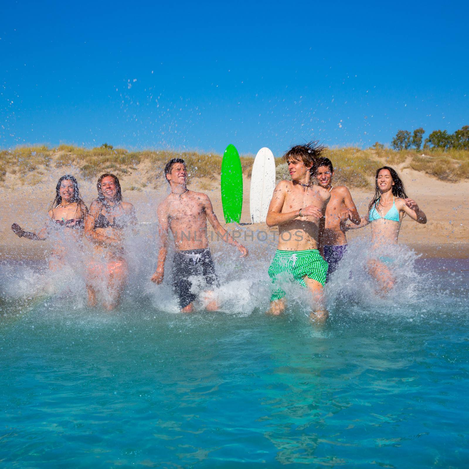 Teen surfers boys and girls group running happy to the beach splashing water