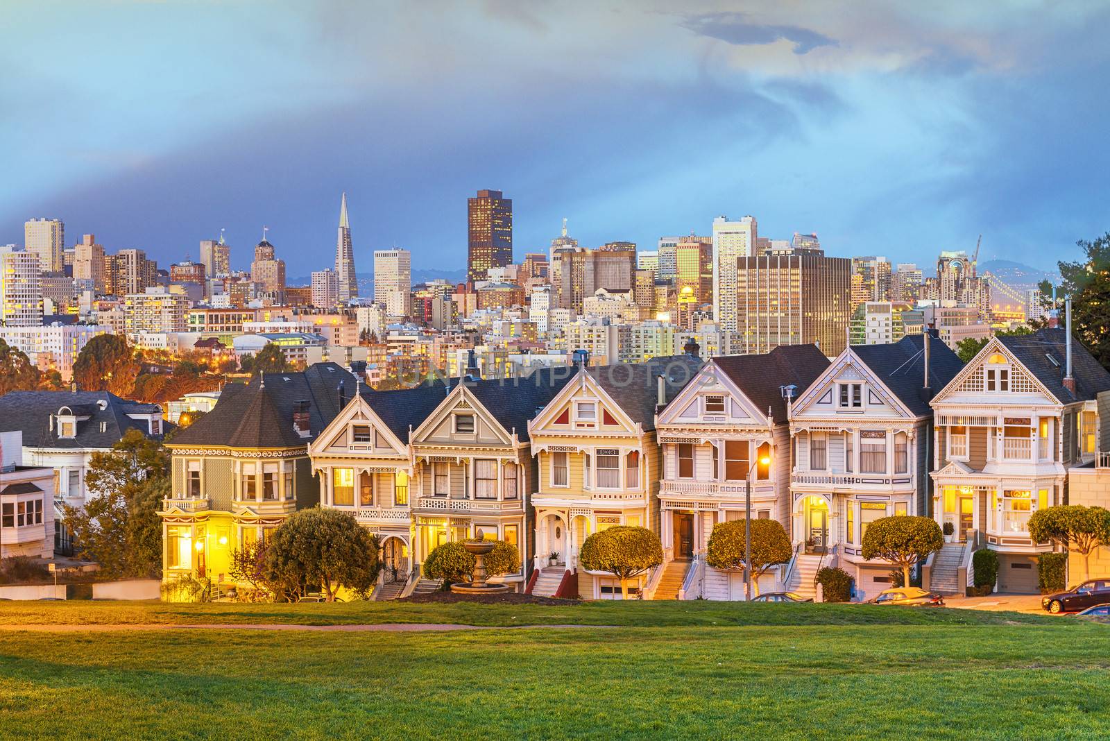 The Painted Ladies of San Francisco, California sit glowing amid the backdrop of a sunset and skyscrapers. 