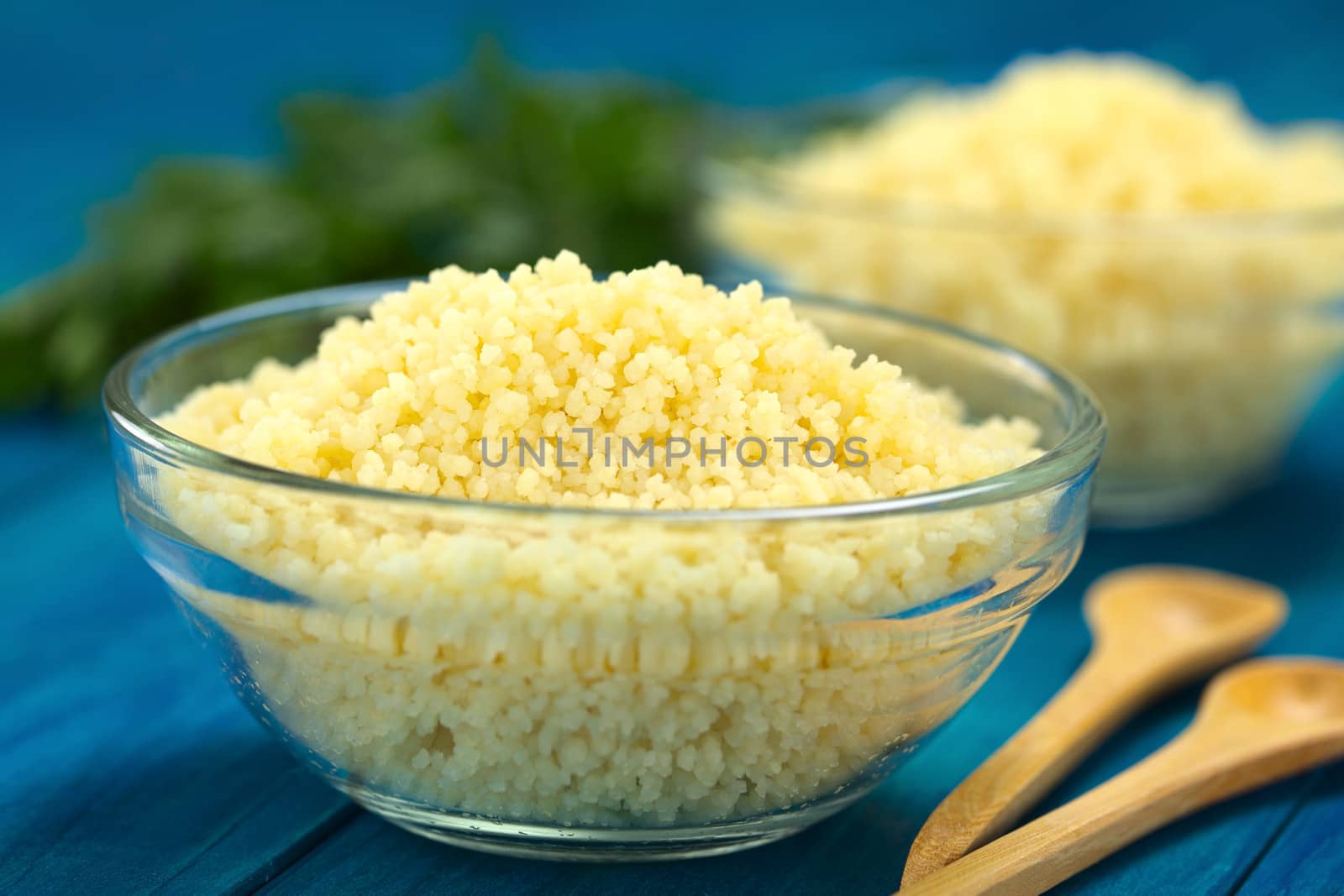 Prepared couscous in glass bowl on blue wooden surface with parsley in the back (Selective Focus, Focus one third into the couscous)