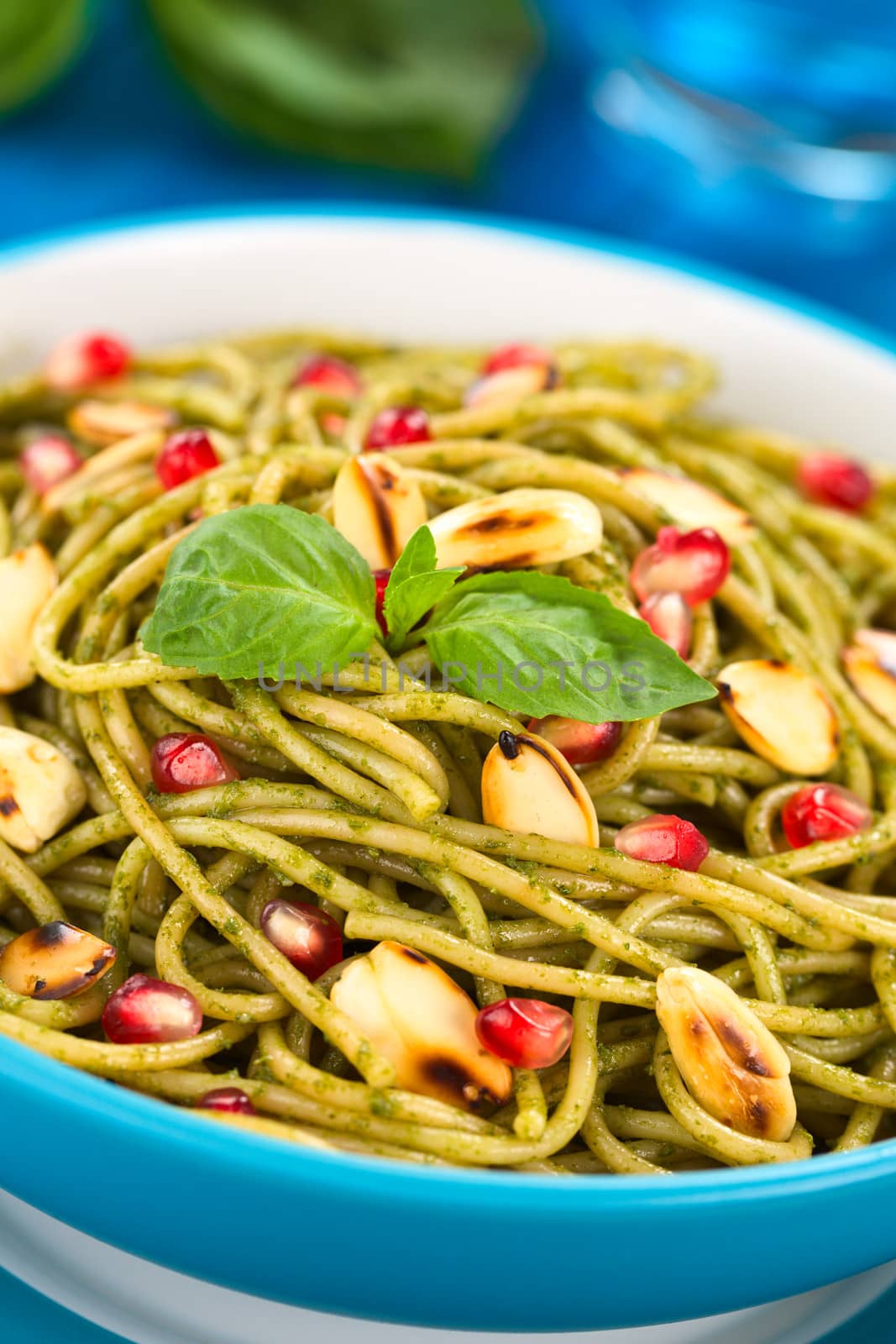 Spaghetti with pesto, pomegranate and roasted almond halves garnished with a basil leaf and served in a blue bowl on blue wood, with glass of water and basil leaf in the back (Selective Focus, Focus on the front of the basil leaf on the dish)