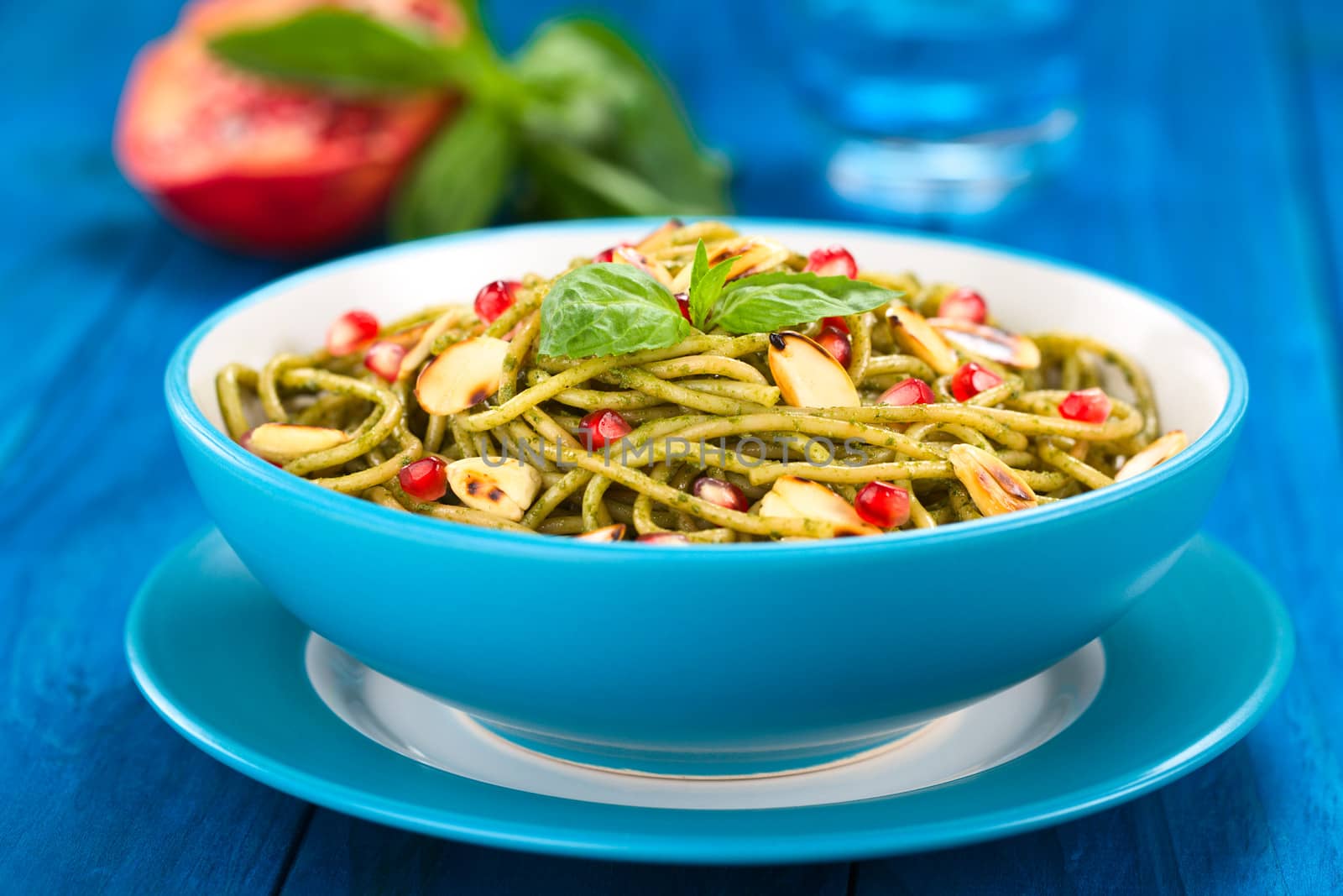 Spaghetti with pesto, pomegranate and roasted almond halves garnished with a basil leaf and served in a blue bowl on blue wood, with glass of water, half pomegranate and basil leaf in the back (Selective Focus, Focus on the front of the basil leaf on the dish)