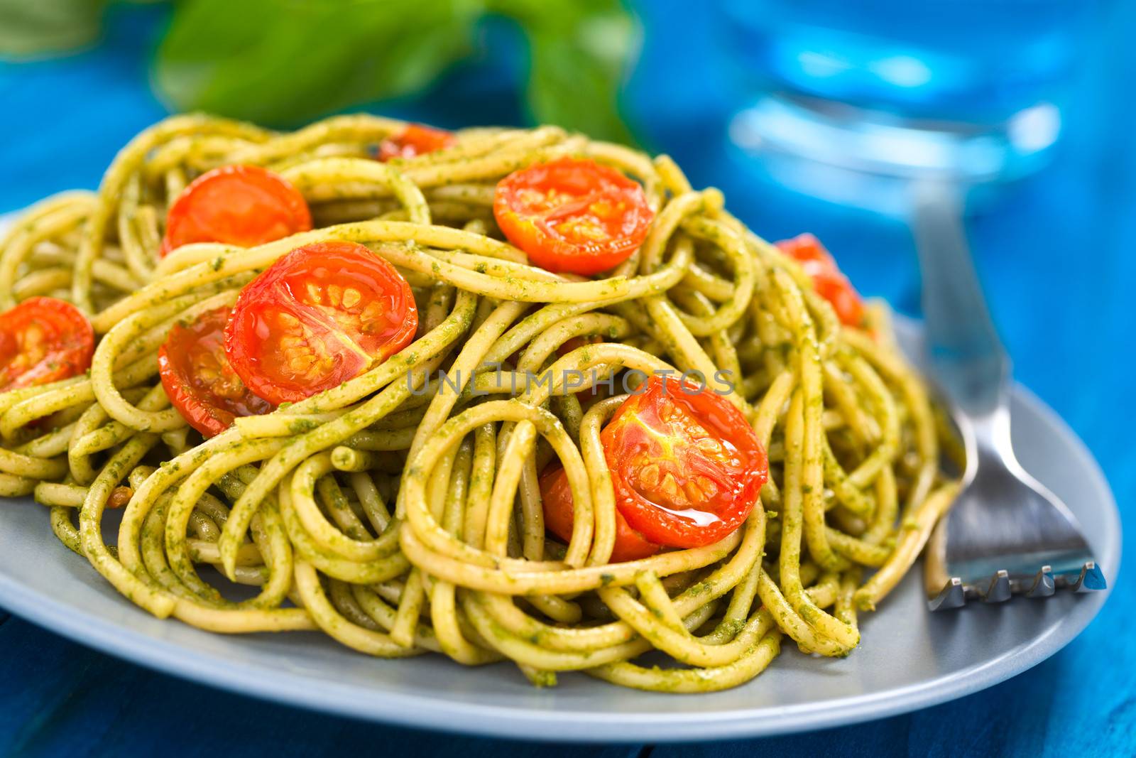 Spaghetti with pesto and baked cherry tomato halves served on a blue plate with a fork beside on blue wood, with a glass of water and basil leaf in the back (Selective Focus, Focus one third into the dish)