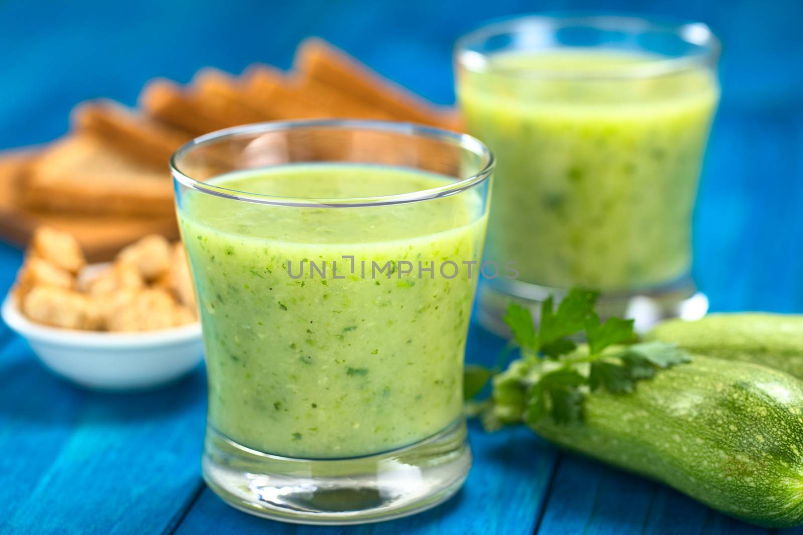 Zucchini cream soup served in glasses on blue wood with small zucchini and parsley on the side, croutons and toast bread in the back (Selective Focus, Focus on the front of the rim of the first glass)