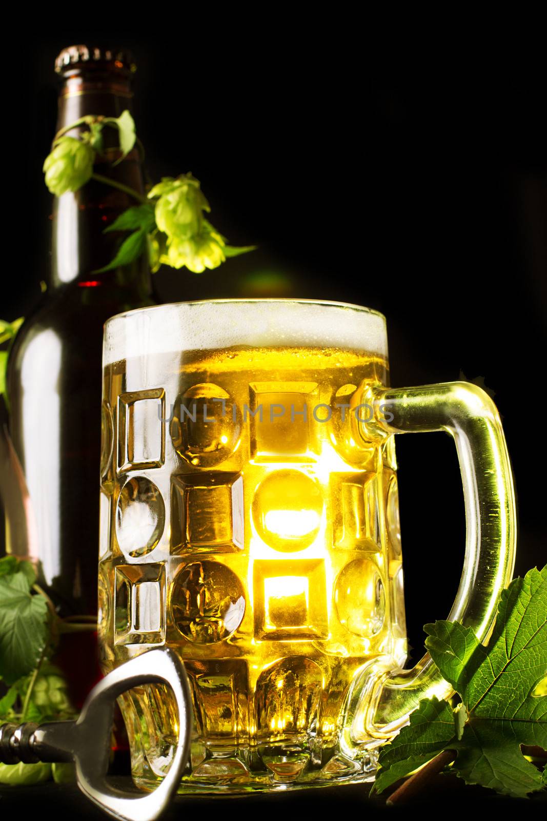 Mug of golden beer, bottle and openner with hop leaves over black