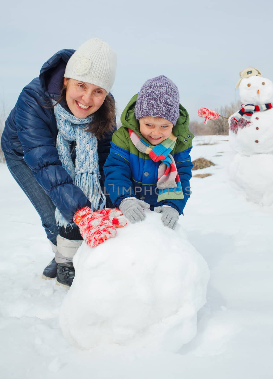 Happy beautiful boy with mother building snowman outside in winter time