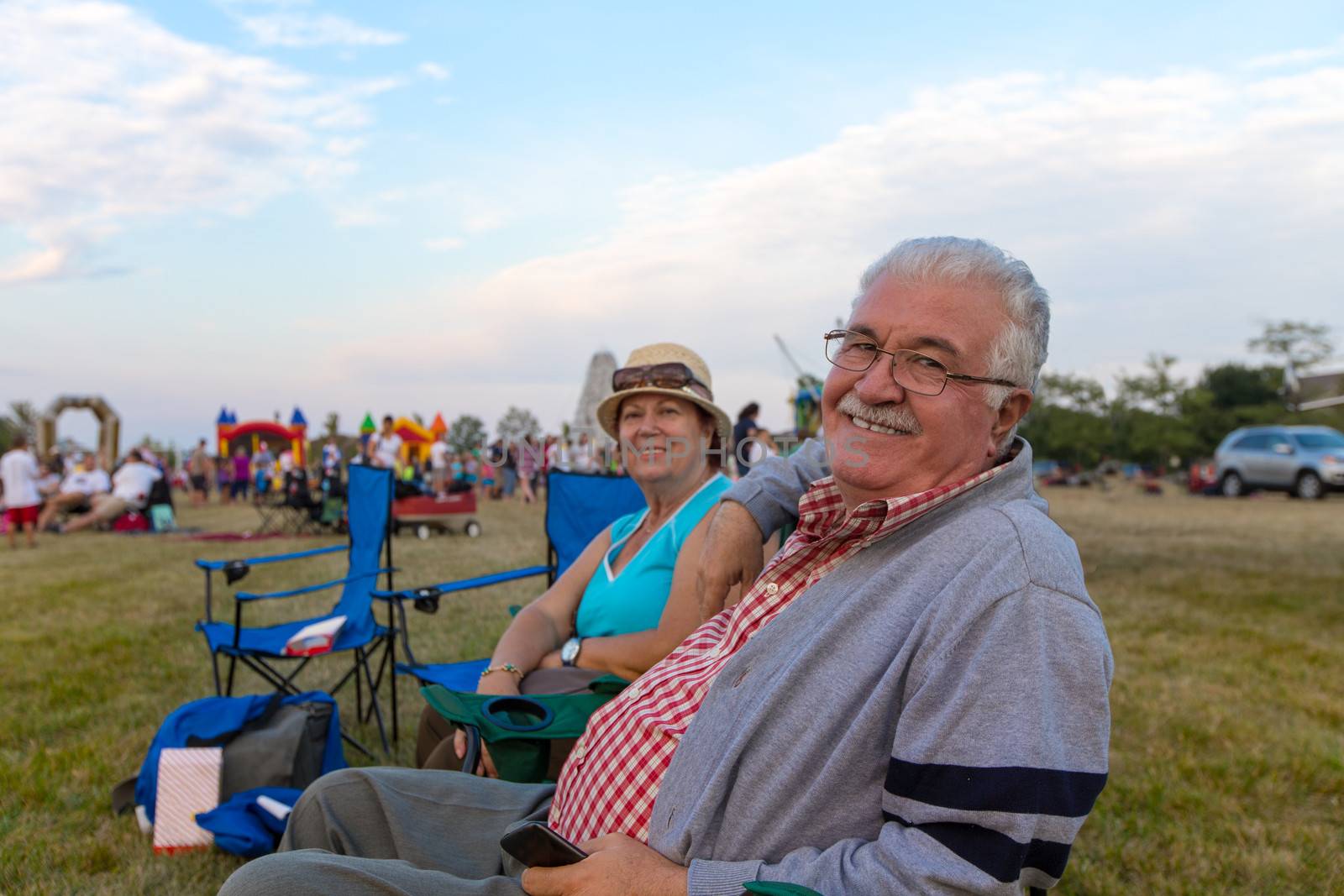 Elderly couple of spectators sitting in deckchairs at an outdoors event on a field turning to smile at the camera