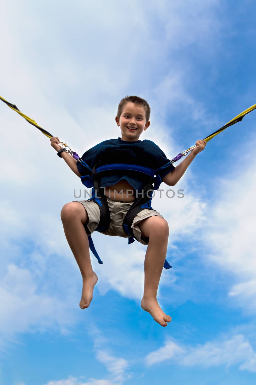 Young boy swinging high in the air against a cloudy blue summer sky in a harness attached to cables or ropes with a beaming smile of enjoyment
