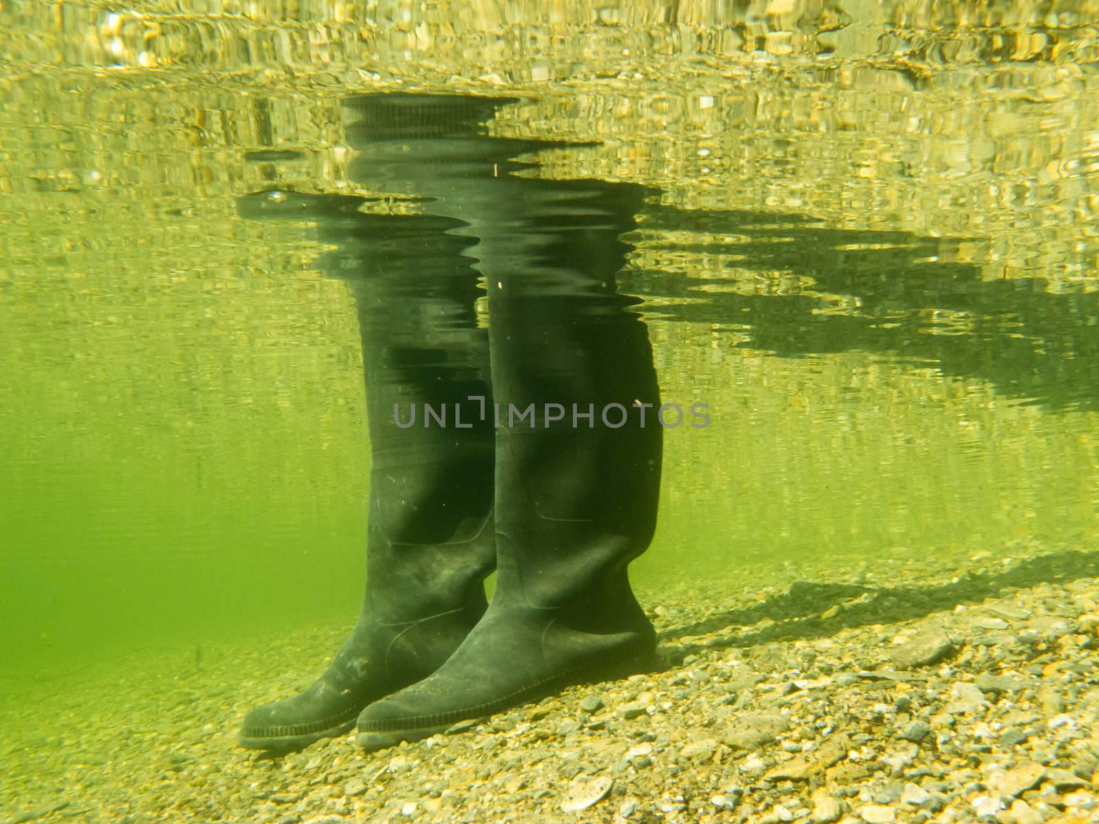 Closeup underwater view of rubber boots gumboots or waders of a person walking in shallow water of gravel and sand beach