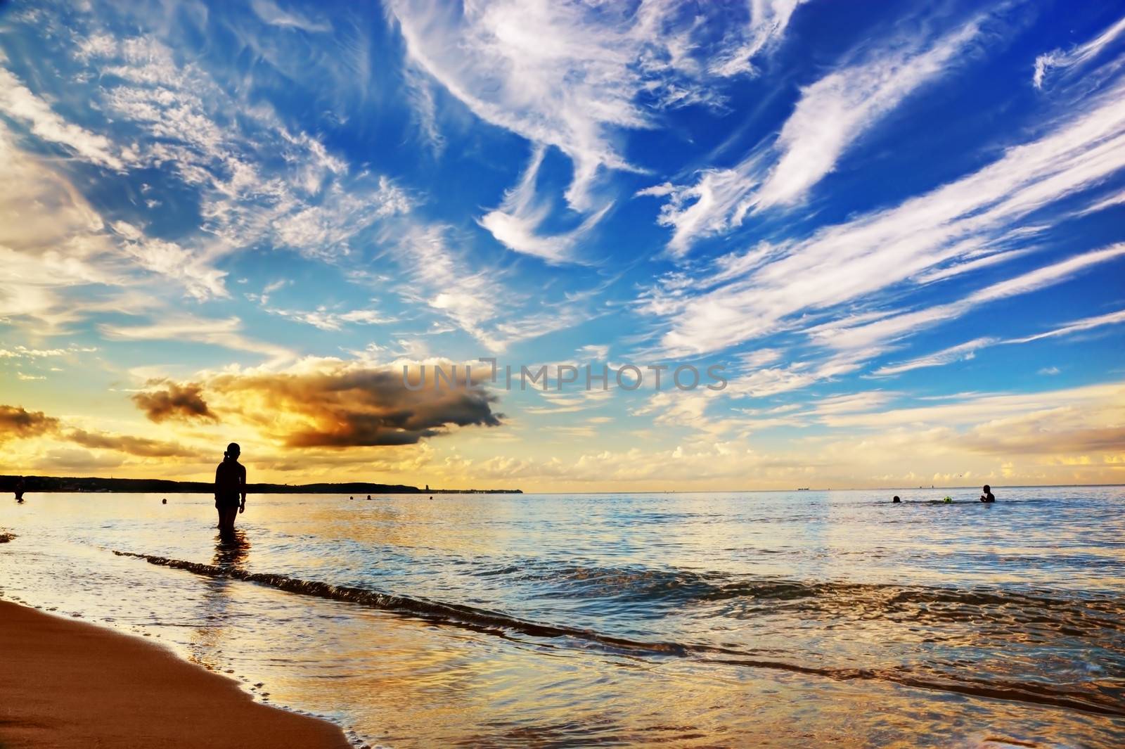 Woman standing in calm ocean under dramatic sunset sky. Summertime