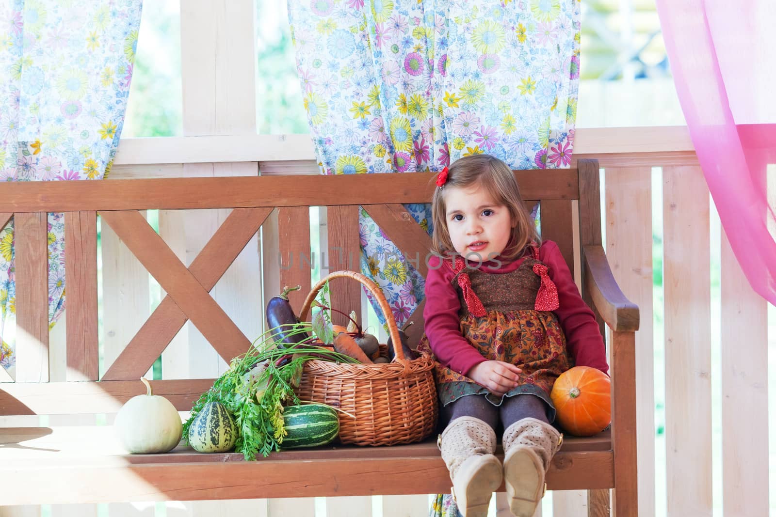 little girl sits on a bench on a terrace with a basket with vegetables