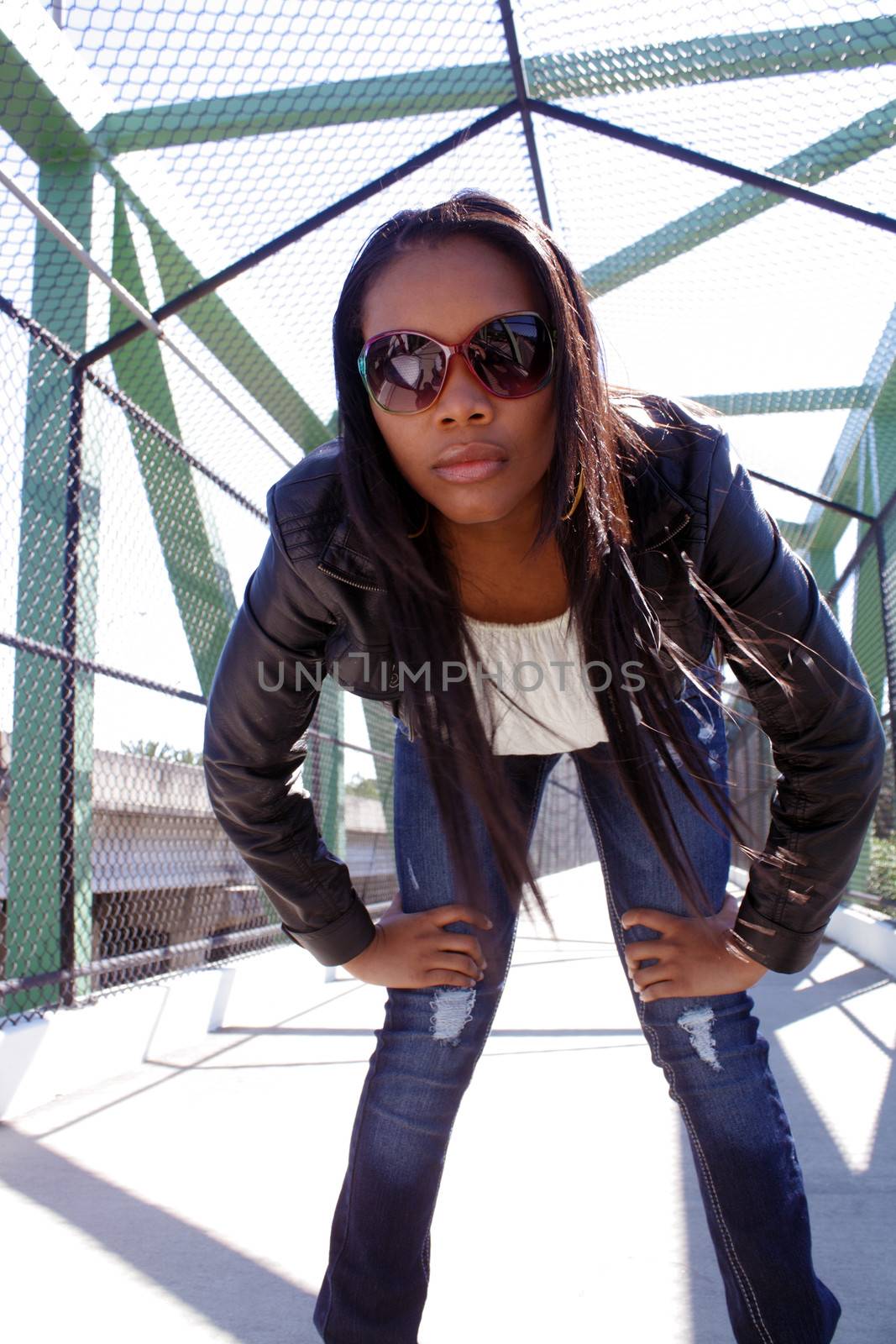 A lovely young Haitian girl on a pedestrian overpass covered with chain-link fence, bends over toward the low-angle camera.