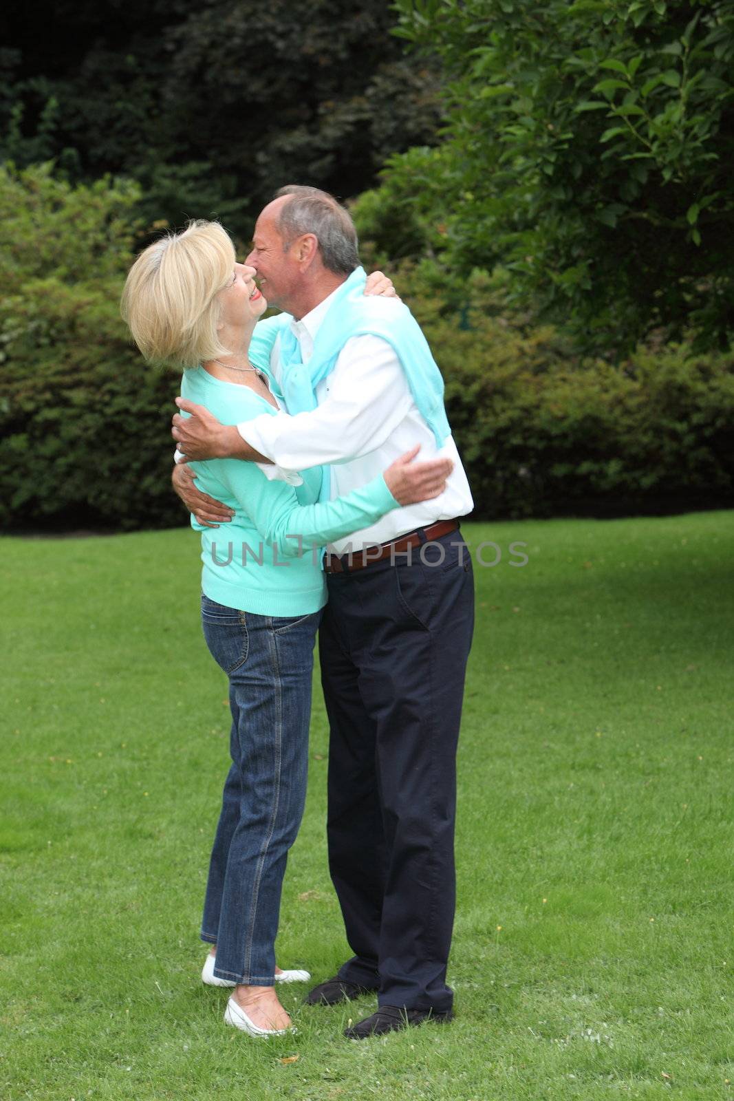 Romantic senior couple hugging each other and kissing as they stand close together in a green summer garden, full length portrait