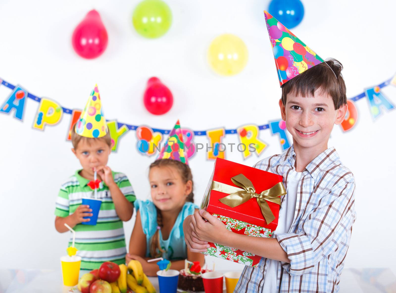 Handsome boy with giftbox looking at camera having fun at birthday party with his friends on background