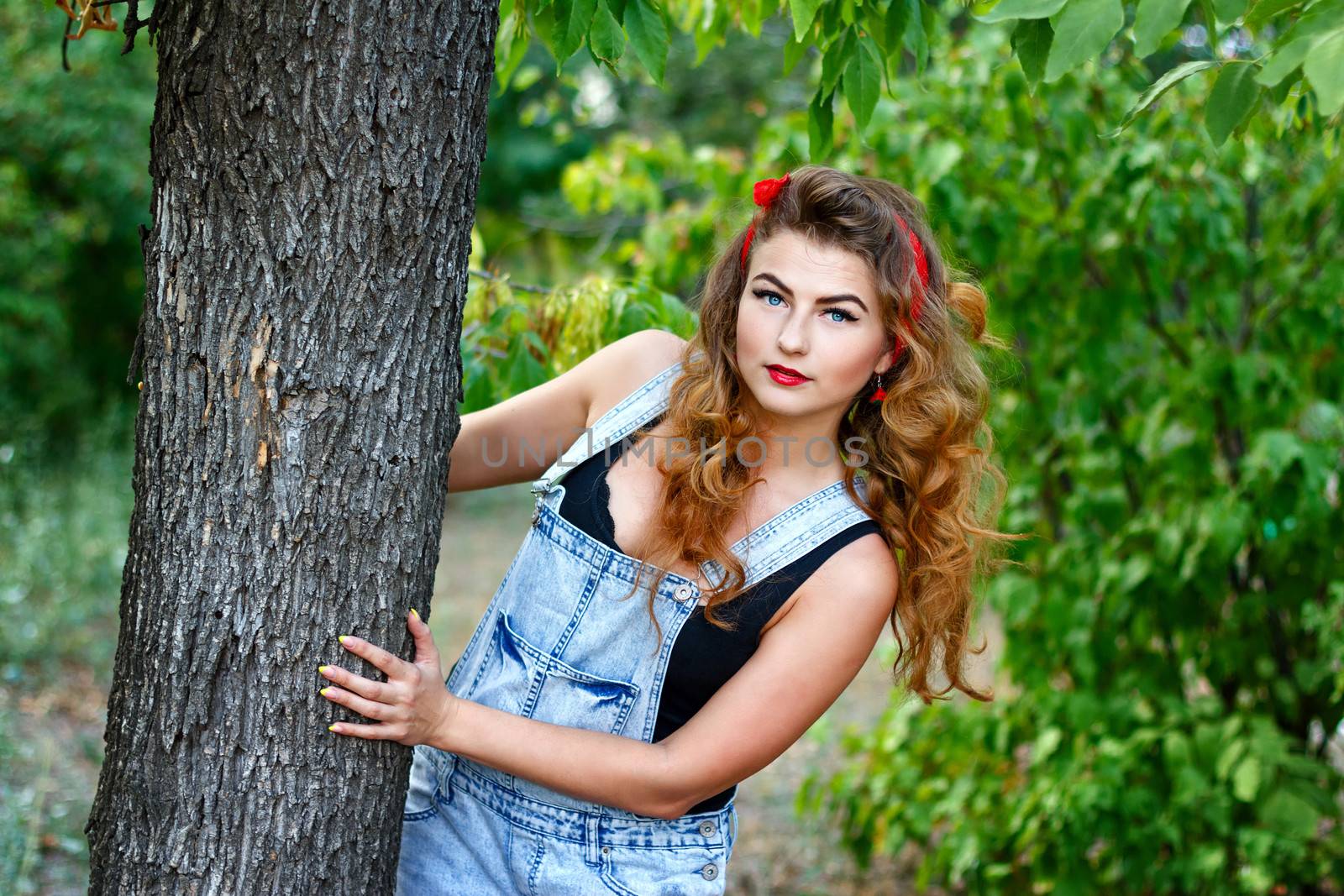 Beautiful pin-up girl in denim overalls and a a red kerchief hiding behind a tree