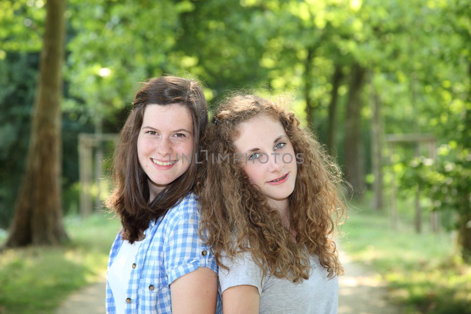 Two pretty healthy teenager girls smiling and posing back to back on an alley in a green forested area, in summer
