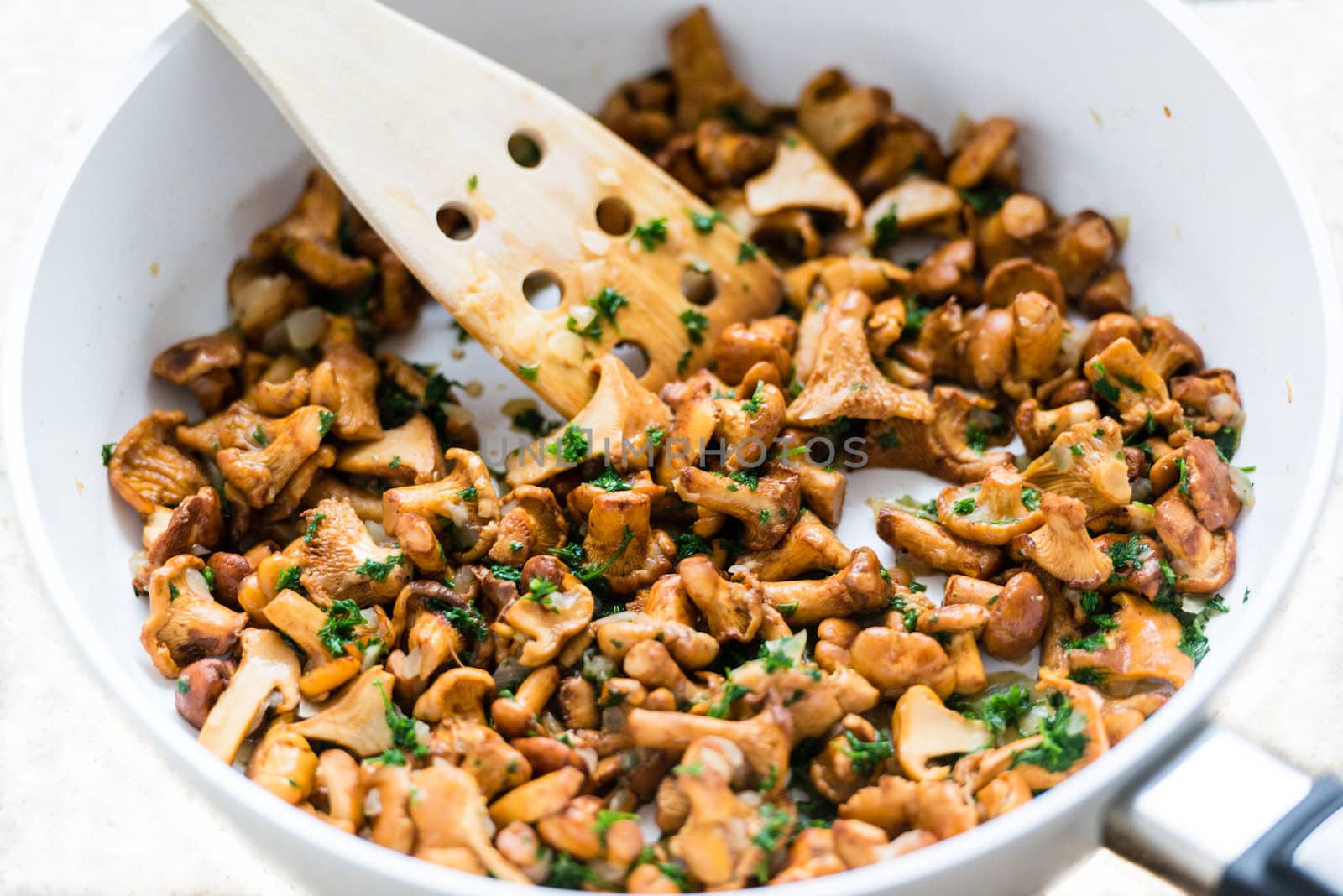 Tasty chanterelles with onion, spices and herbs in a modern white pan with wooden spatula on bright background