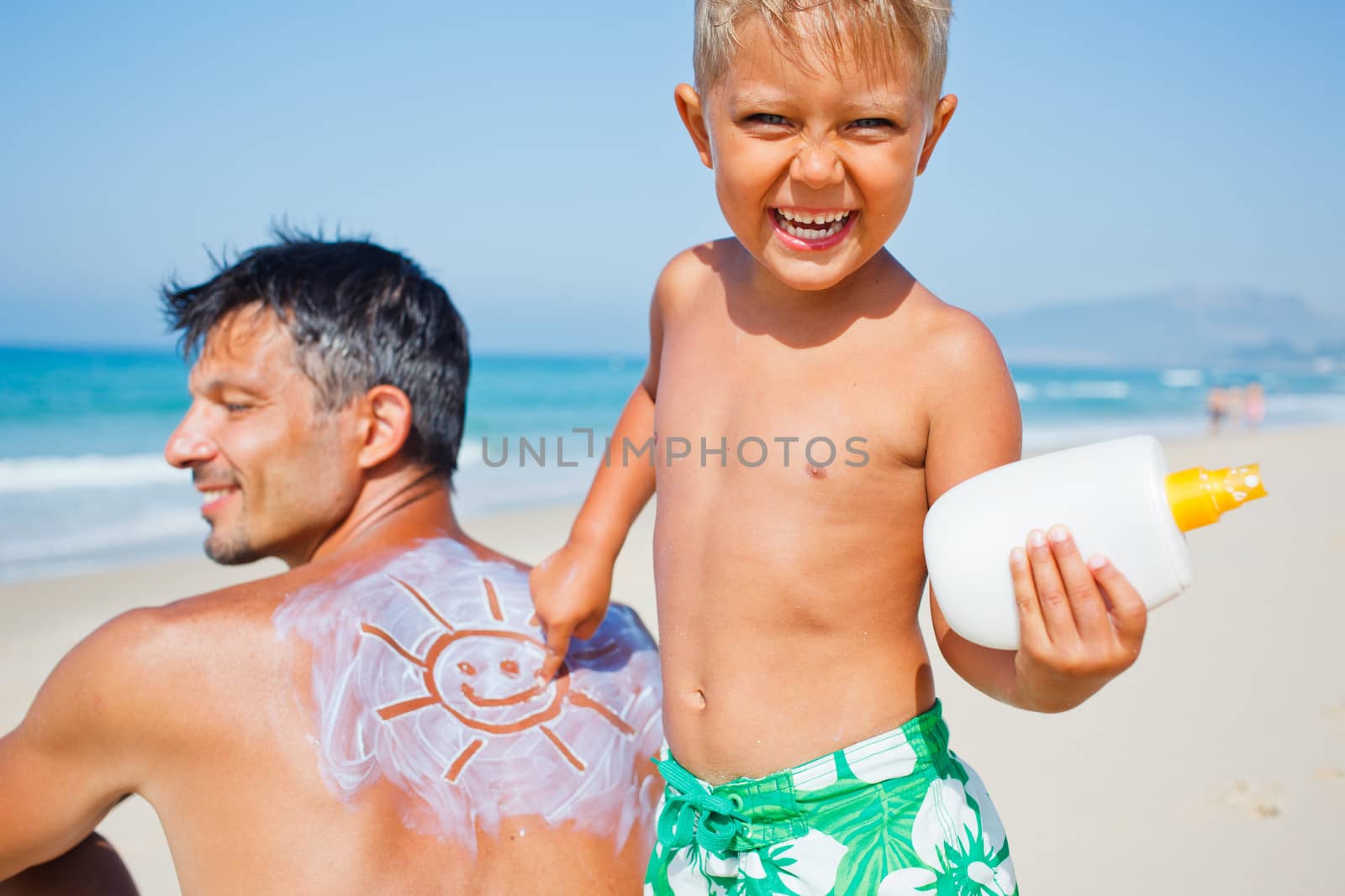 Adorable boy at tropical beach applying sunblock cream on a father's back.