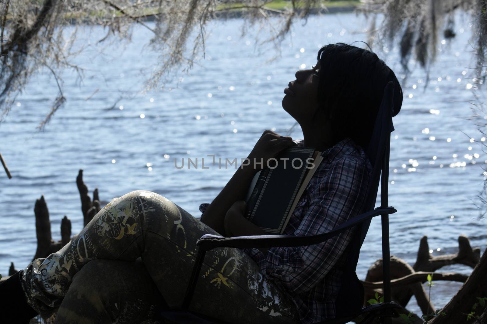 A lovely young woman looks away from her book at the waterfront, in silhouette.