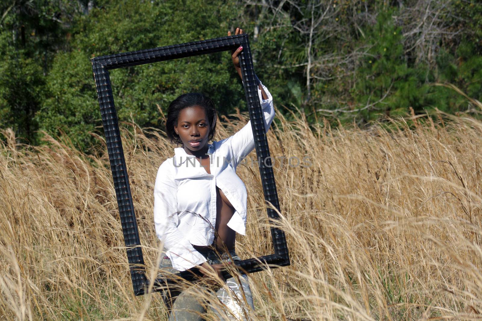 A lovely young black woman outdoors, standing in tall grass, holding a large black picture frame.  Generous copyspace on frame right.