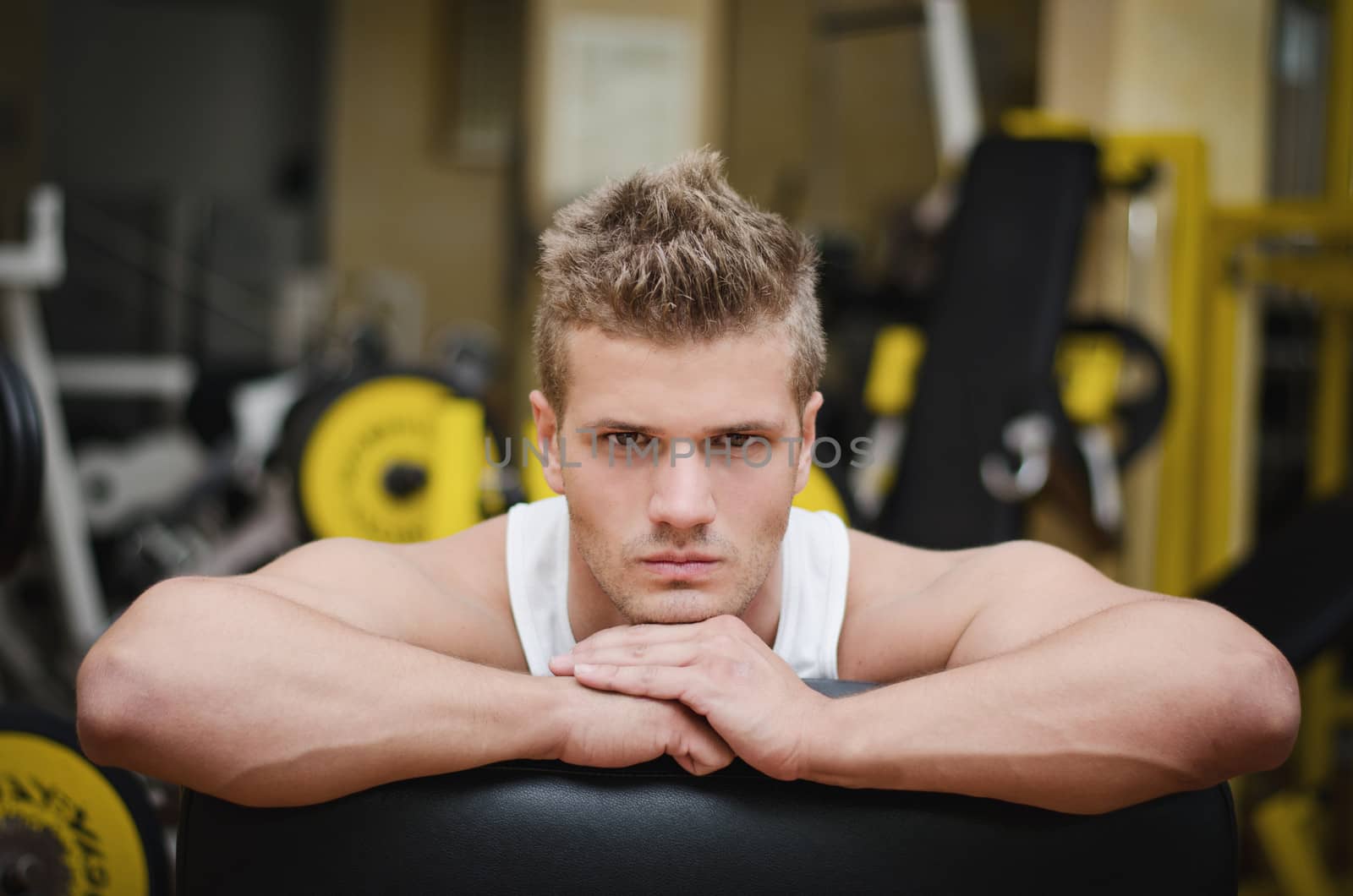 Handsome young man in gym resting on gym equipment, looking in camera