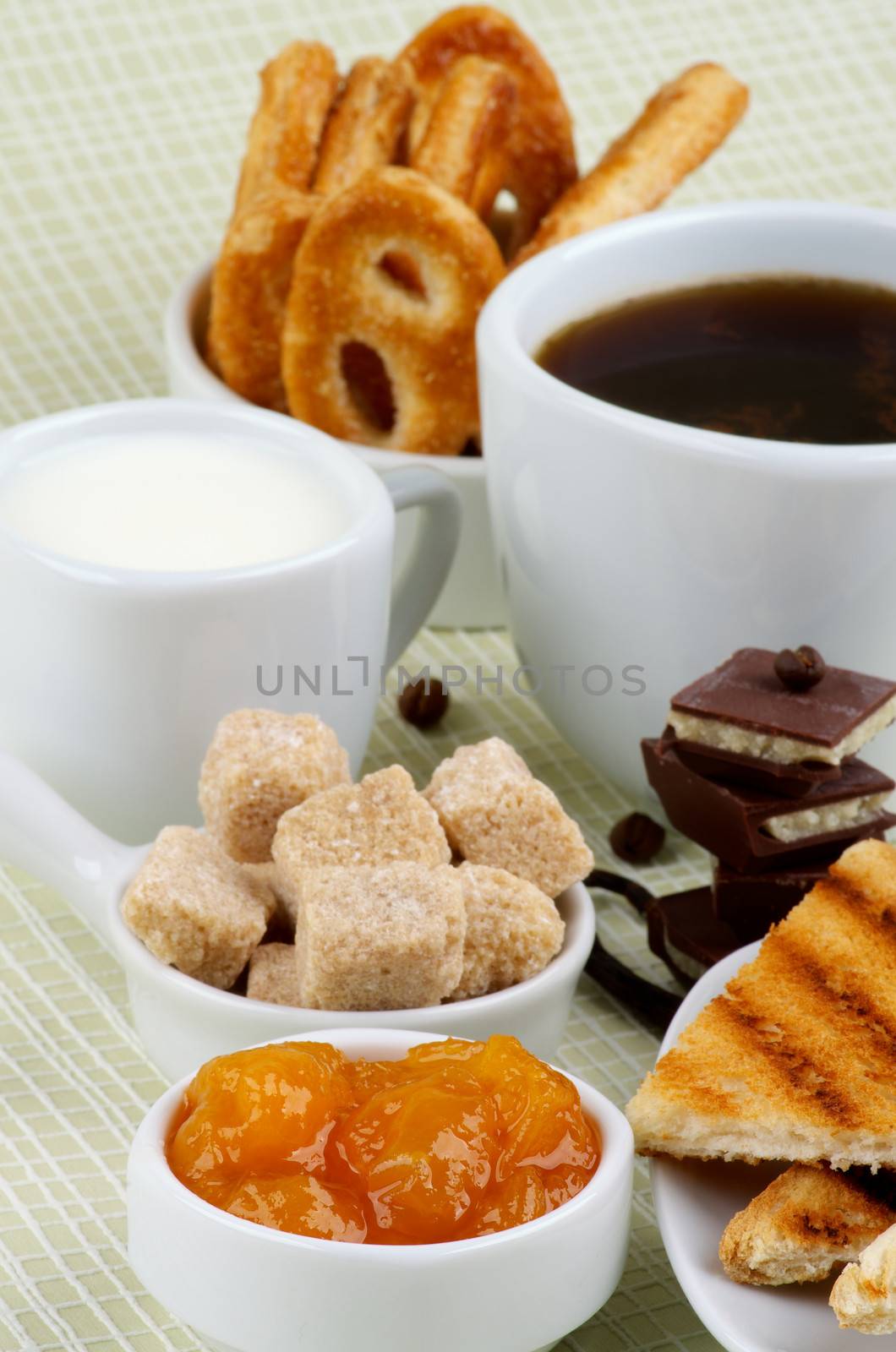 Arrangement of Coffee Cup, Milk, Puff Pastry, Apricot Jam and Toasts with Chocolate and Brown Sugar Cubes closeup on light green Checkered background
