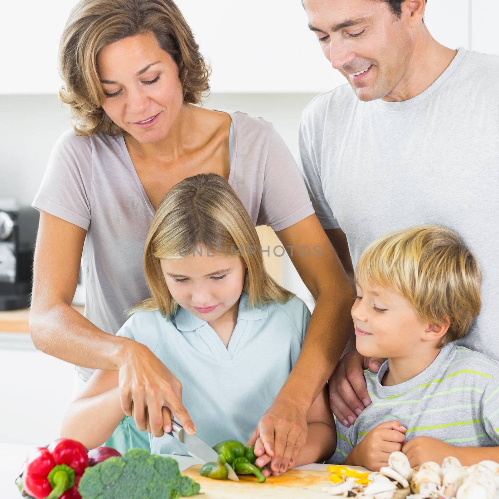 Mother teaching daughter to slice vegetables as father and son are watching in the kitchen