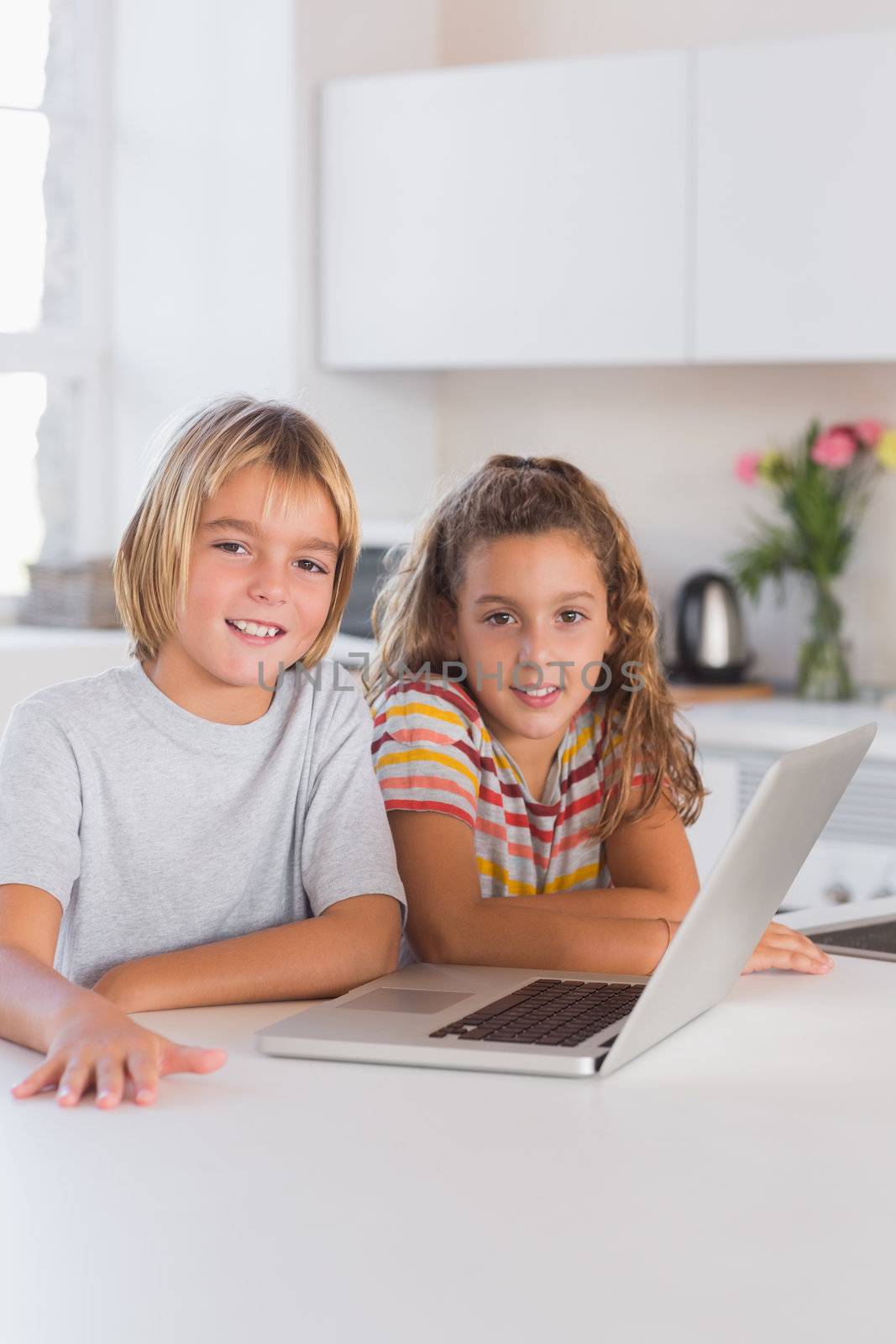 Two children looking at the camera with laptop in front in kitchen