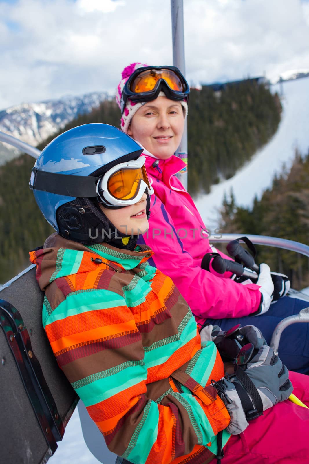 Portrait of happy girl in ski goggles and a helmet with his mother on the ski lift