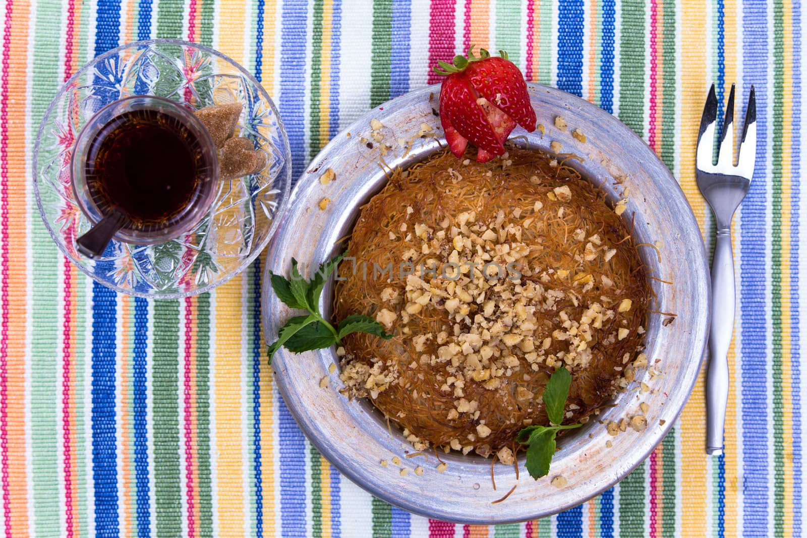 Turkish dessert kunefe on a picnic cloth with mint leaves and sliced strawberry along with Turkish tea and fork