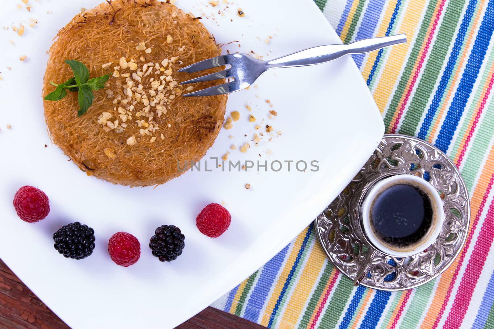 Turkish dessert kunefe  served with Turkish Coffee on a white plate with mint leaves along red and black berries on a picnic table