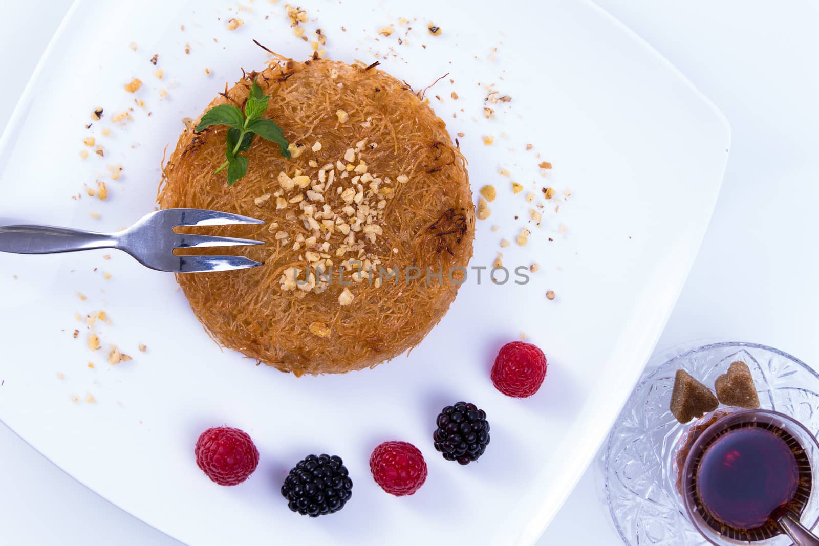 Turkish dessert kunefe  served on a white plate with mint leaves, red and black berries along with Turkish Hot tea