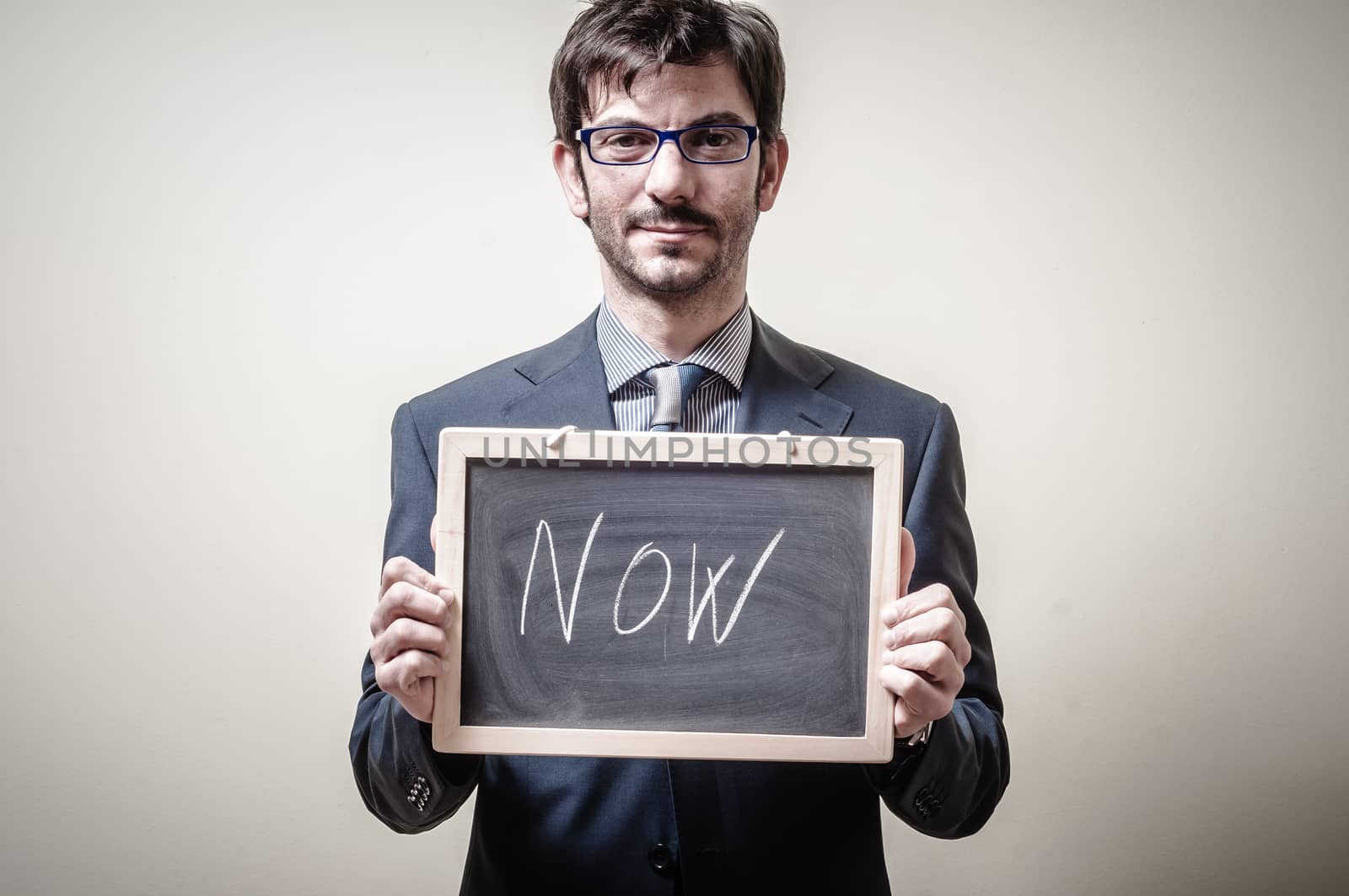businessman holding blackboard written now on gray background