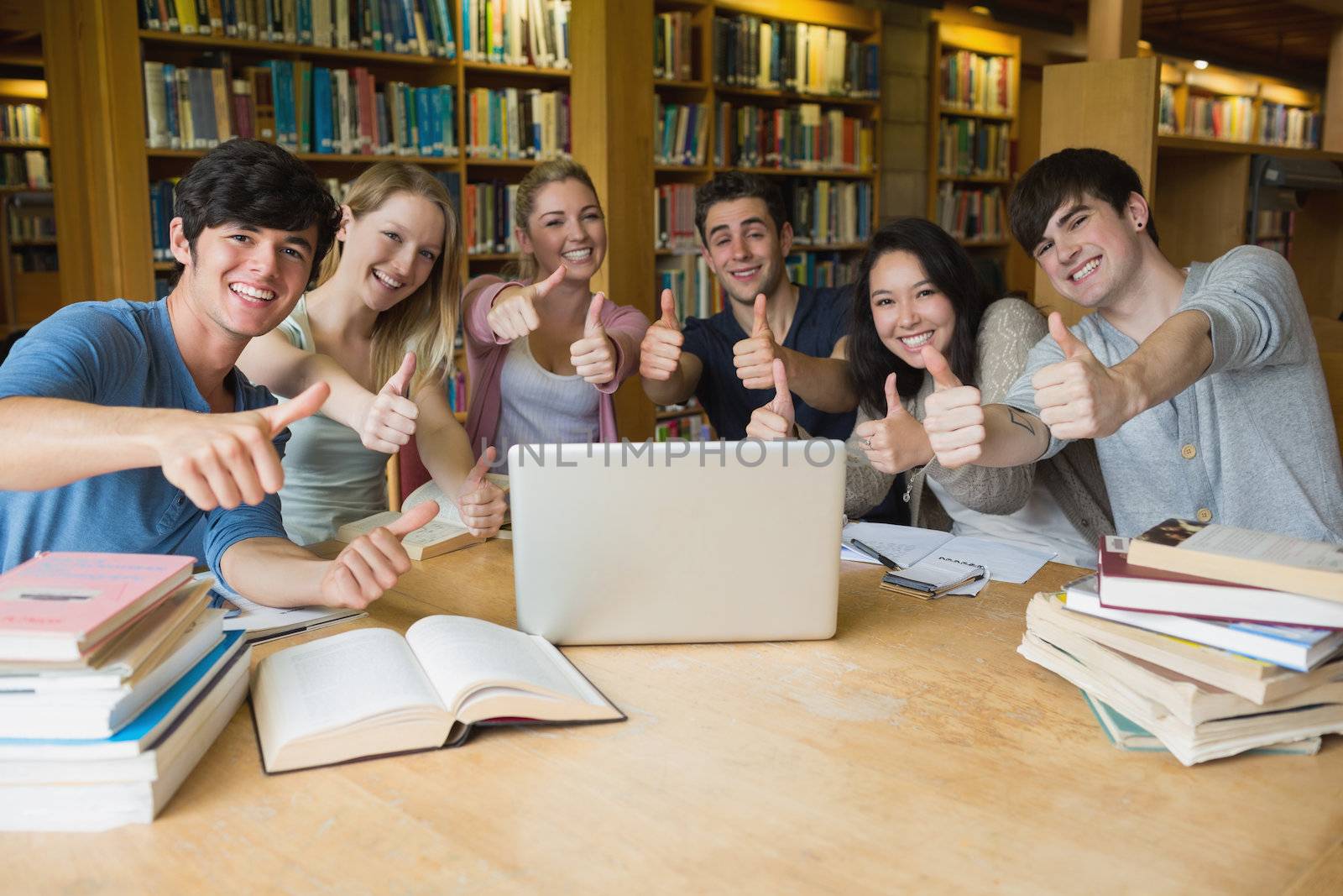 Group of students sitting in a library at a table while using the laptop and giving thumbs up