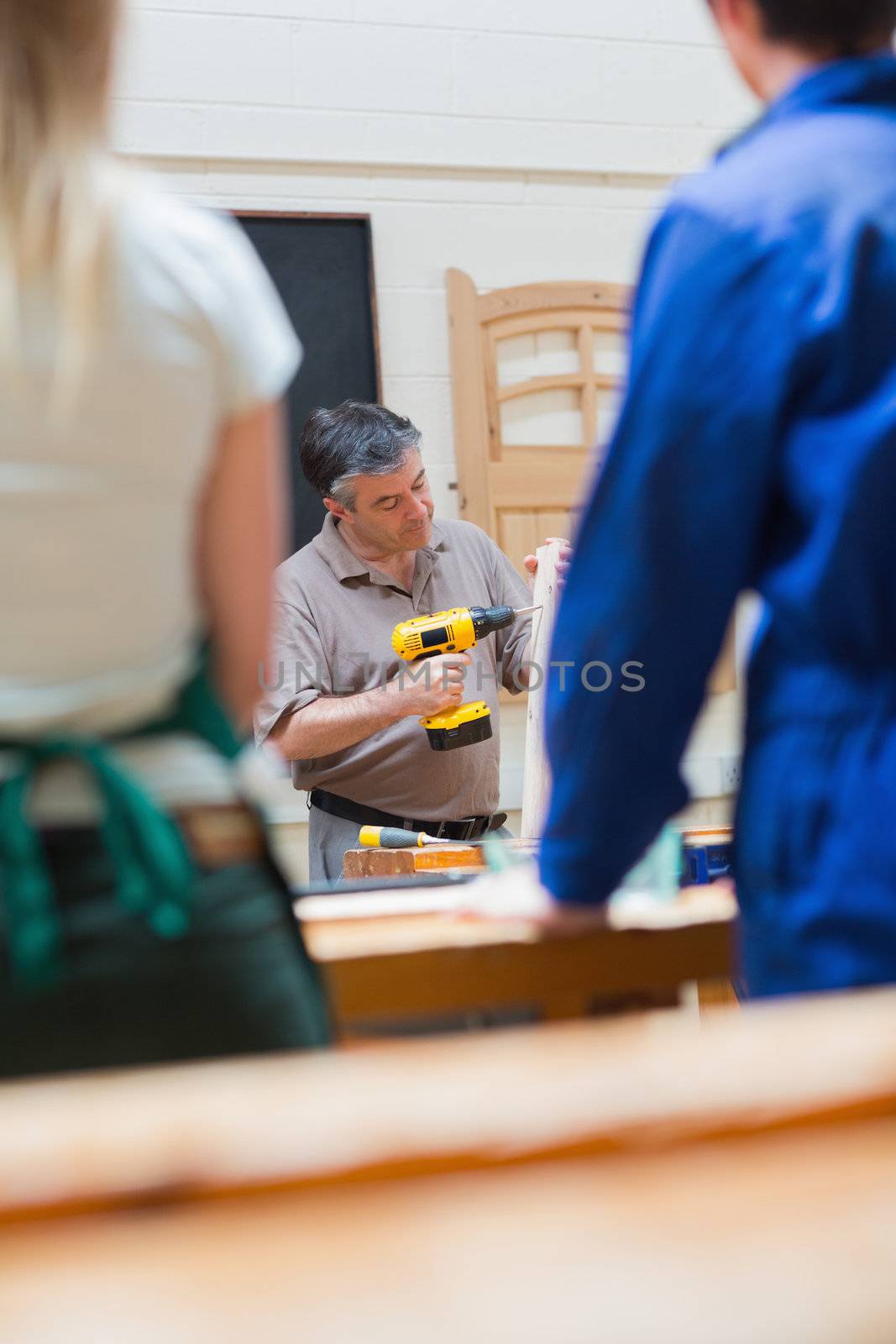 Teacher in a woodwork class holding a wooden board while explaining and drilling