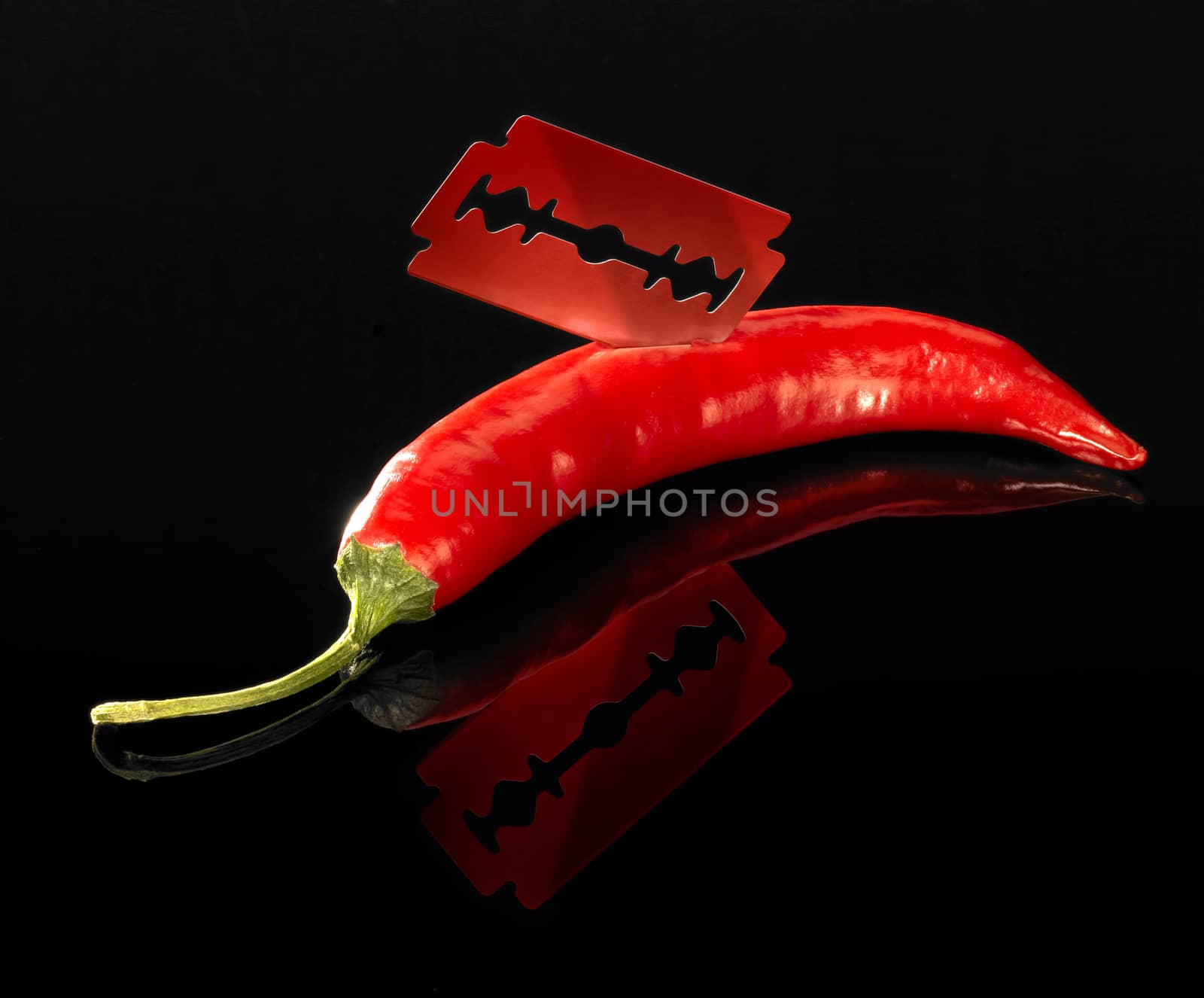 studio shot of a sharp razor blade and red hot chili in dark reflective back