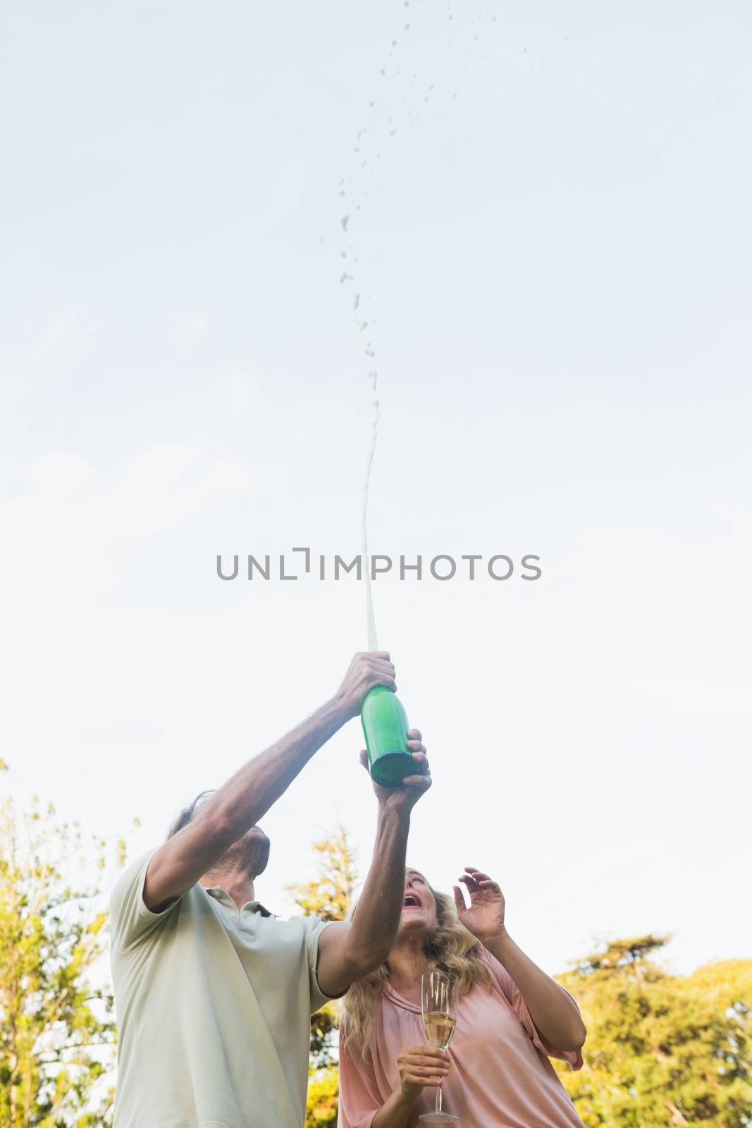 Handsome man spraying bottle of champagne with blonde partner outside in the sunshine