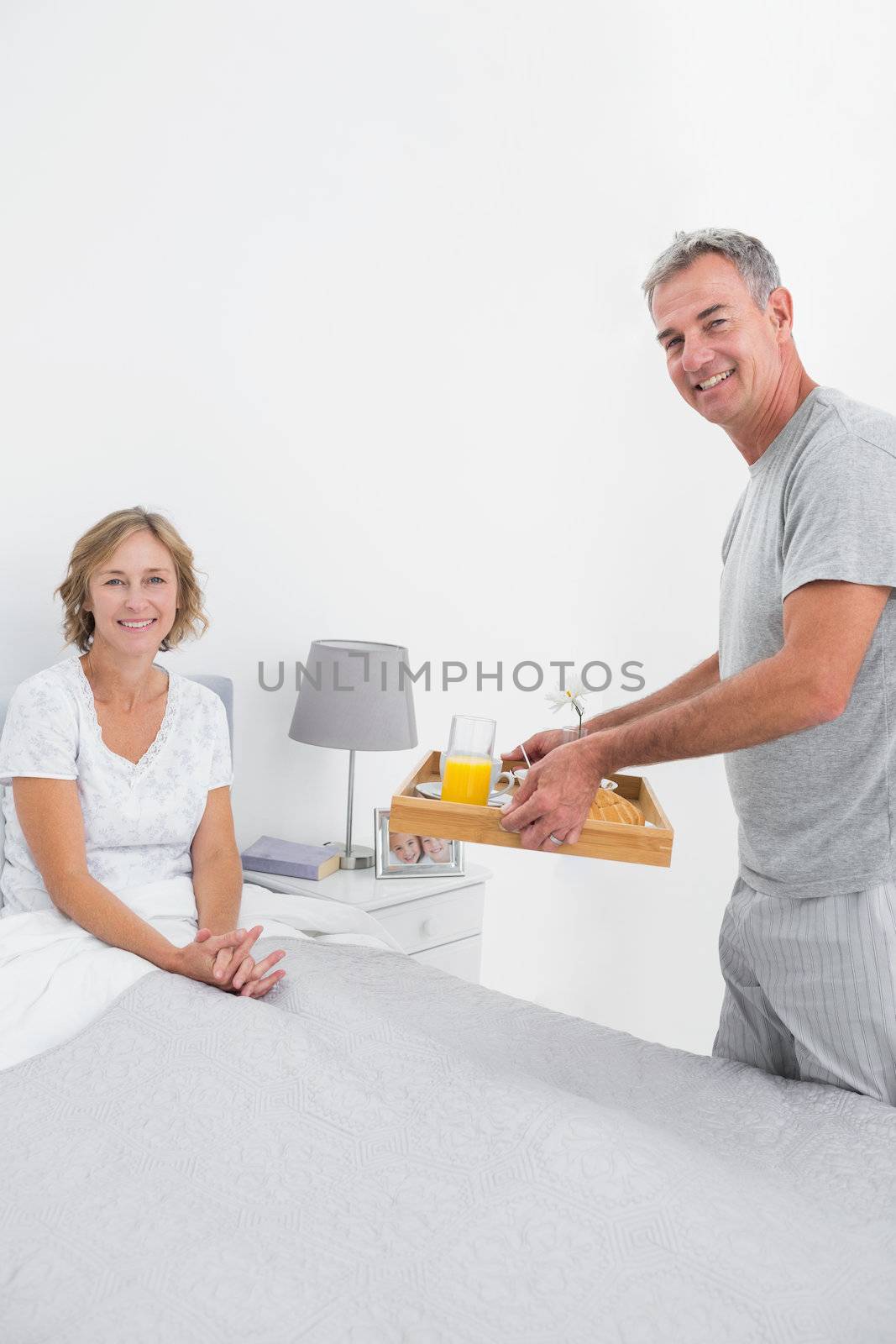 Smiling husband bringing breakfast in bed to wife looking at camera in bedroom at home