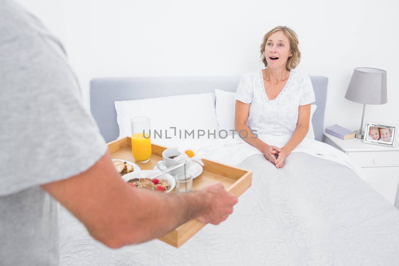 Husband bringing breakfast in bed to surprised wife in bedroom at home