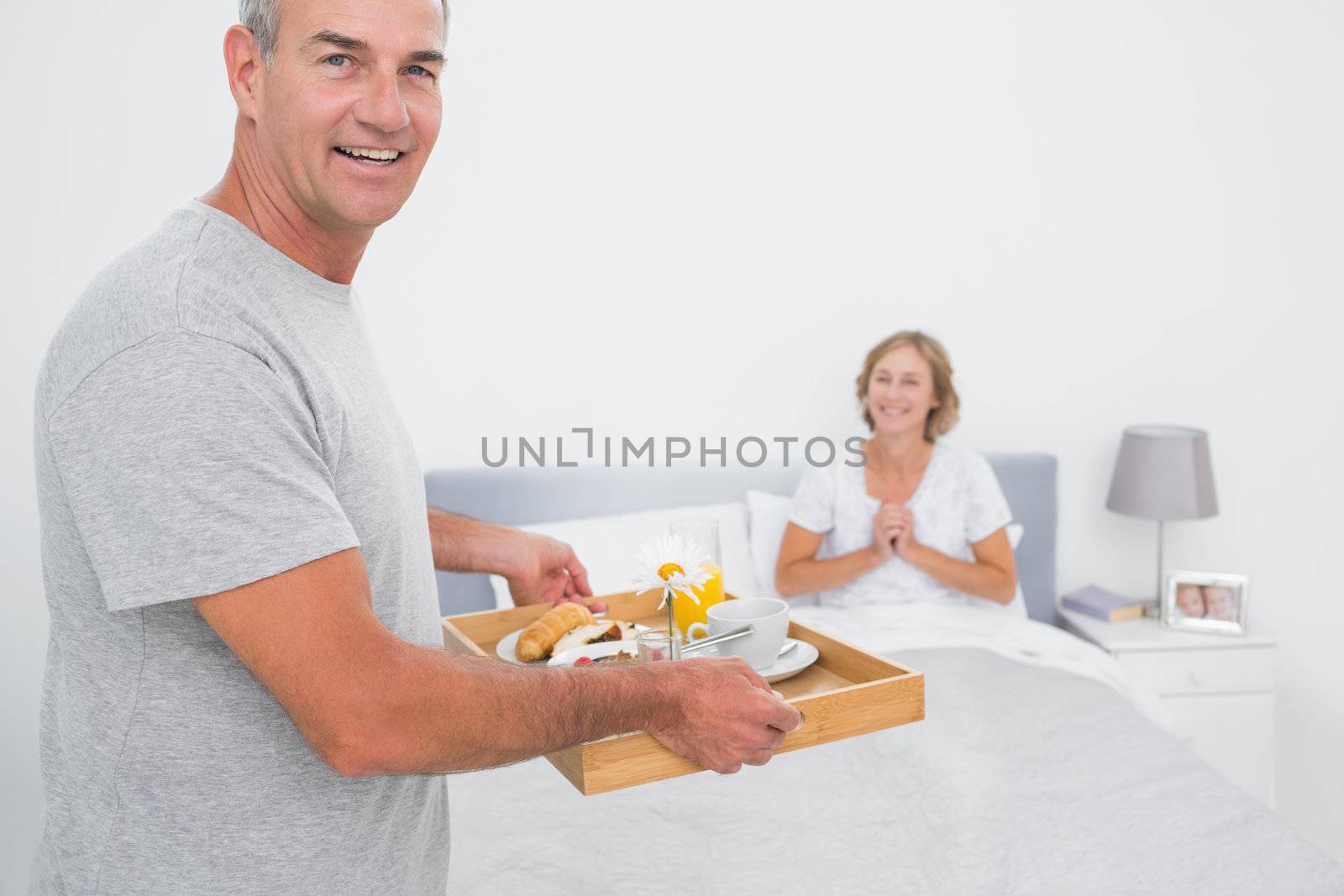 Happy husband bringing breakfast in bed to delighted wife looking at camera in bedroom at home