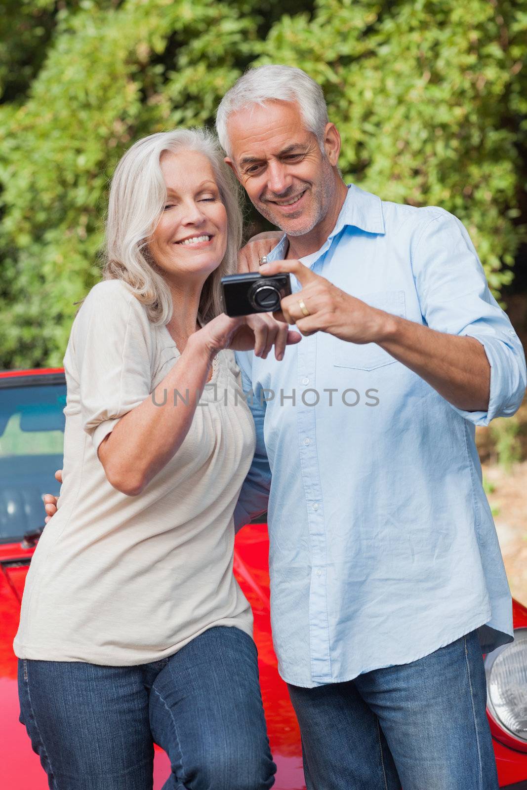 Smiling mature couple looking at pictures on their camera while leaning against their cabriolet