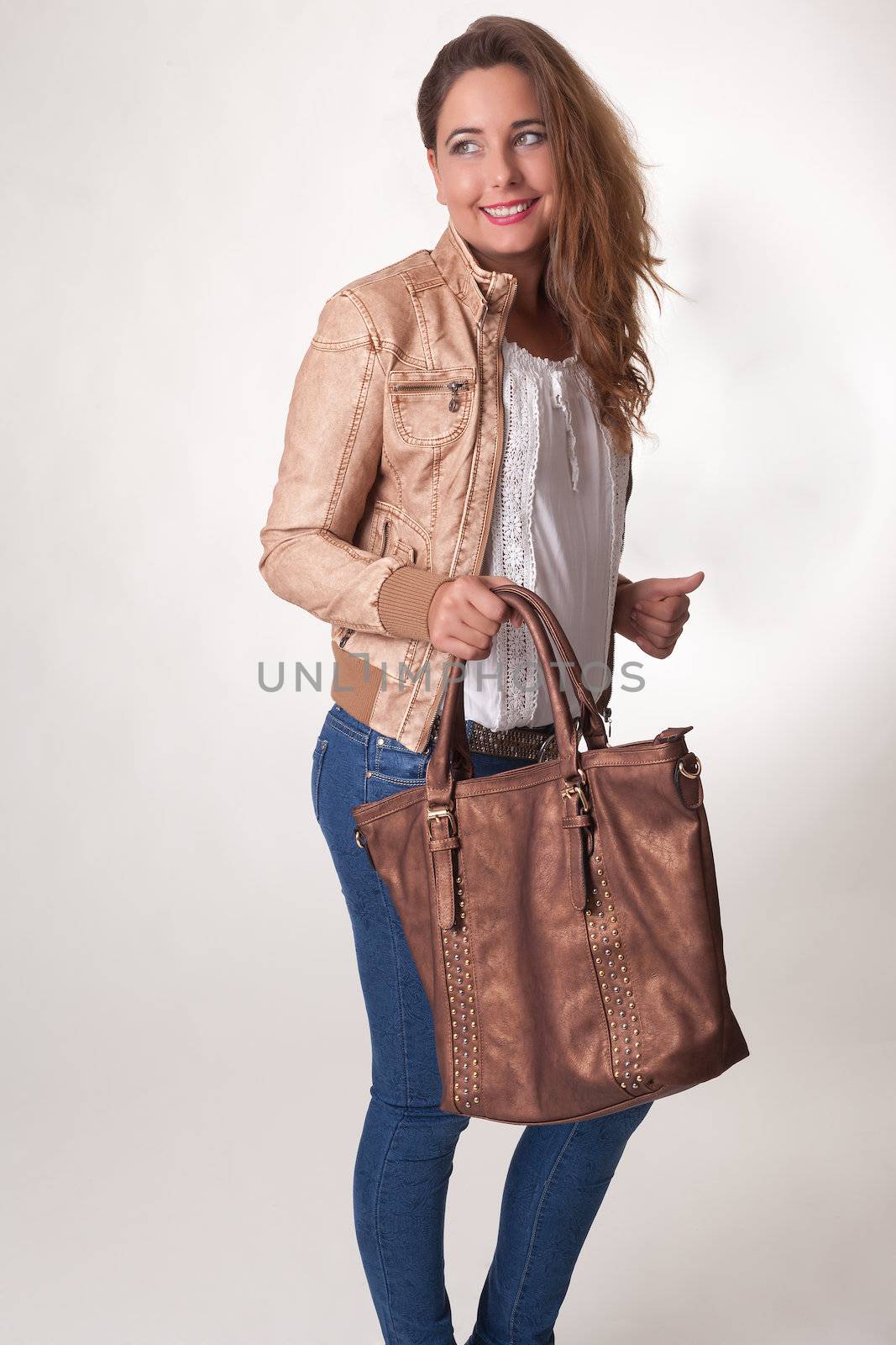 Stylish beautiful young woman with a large brown leather handbag in her hand standing looking back over her shoulder with a friendly smile, studio portrait