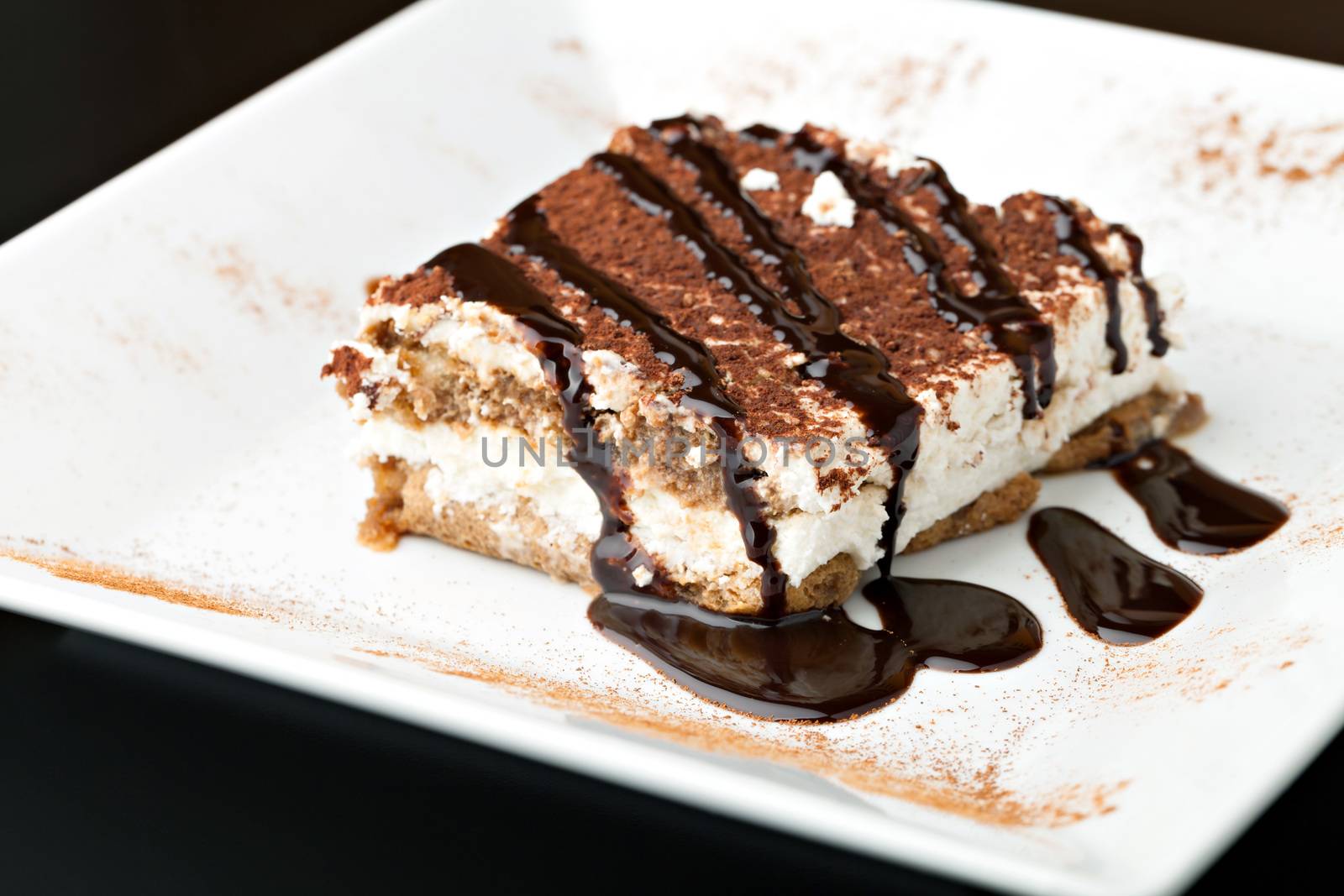 Slice of delicious tirimisu cake on a white plate garnished with cocoa power and drizzled chocolate sauce.
