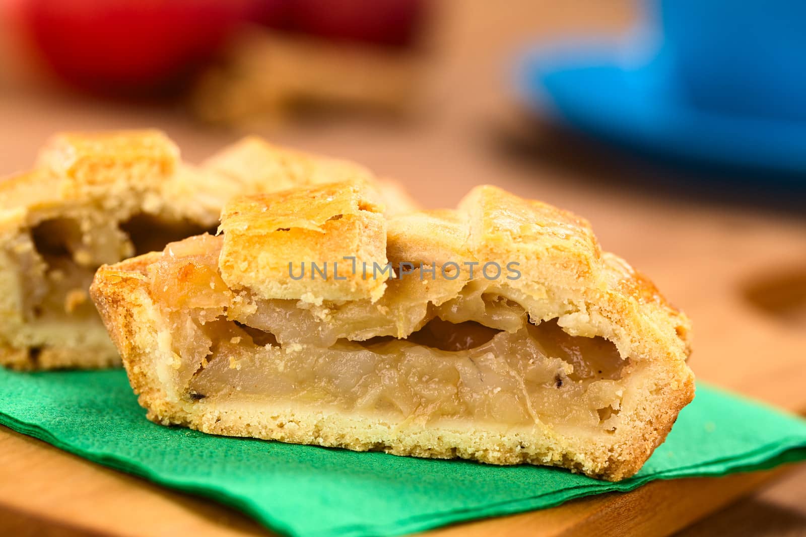 Half apple pie with lattice crust on green napkin on wooden board, with a blue cup, apples and cinnamon sticks in the back  (Selective Focus, Focus on the front of the filling of the pie) 